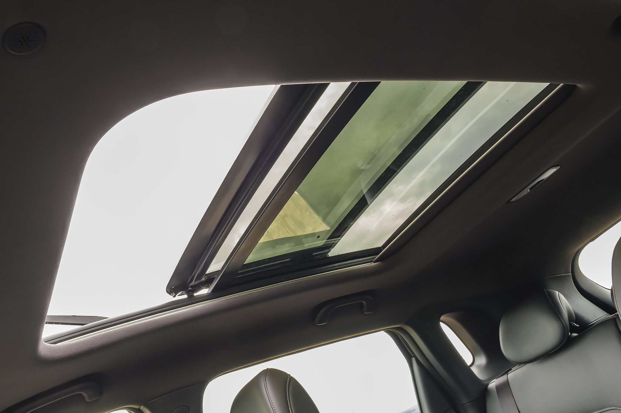 Teto solar panorâmico de um carro, com duas seções de vidro, uma aberta e outra fechada, revelando o céu claro e reflexos esverdeados. O interior do veículo é escuro, com parte do banco de couro visível.