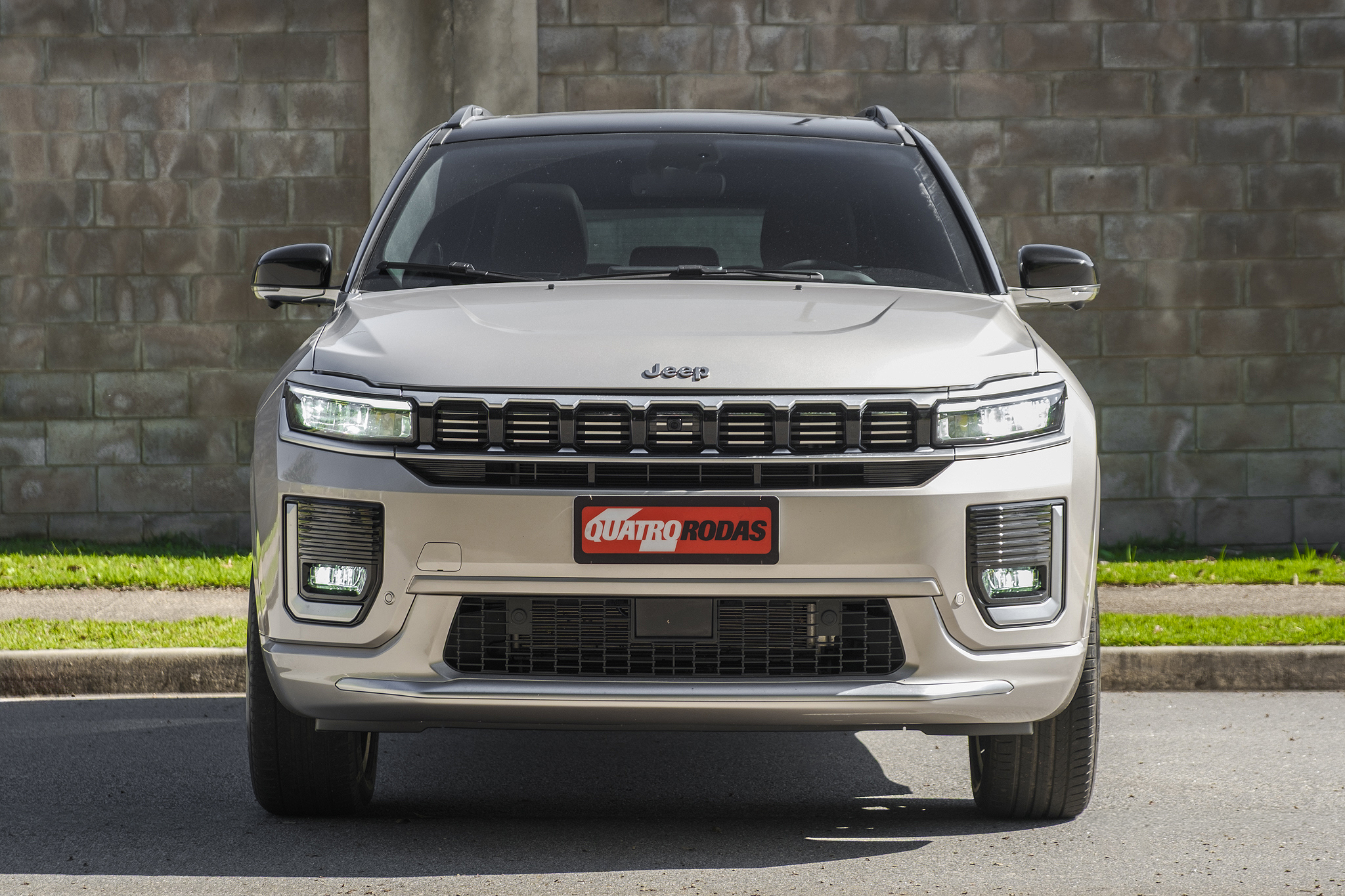 SUV Jeep Commander bege claro, visto de frente, com faróis e luzes de neblina acesos, grade frontal preta e cromada, e placa vermelha Quatro Rodas no para-choque. O veículo está estacionado em uma rua asfaltada, com uma parede de tijolos e grama ao fundo
