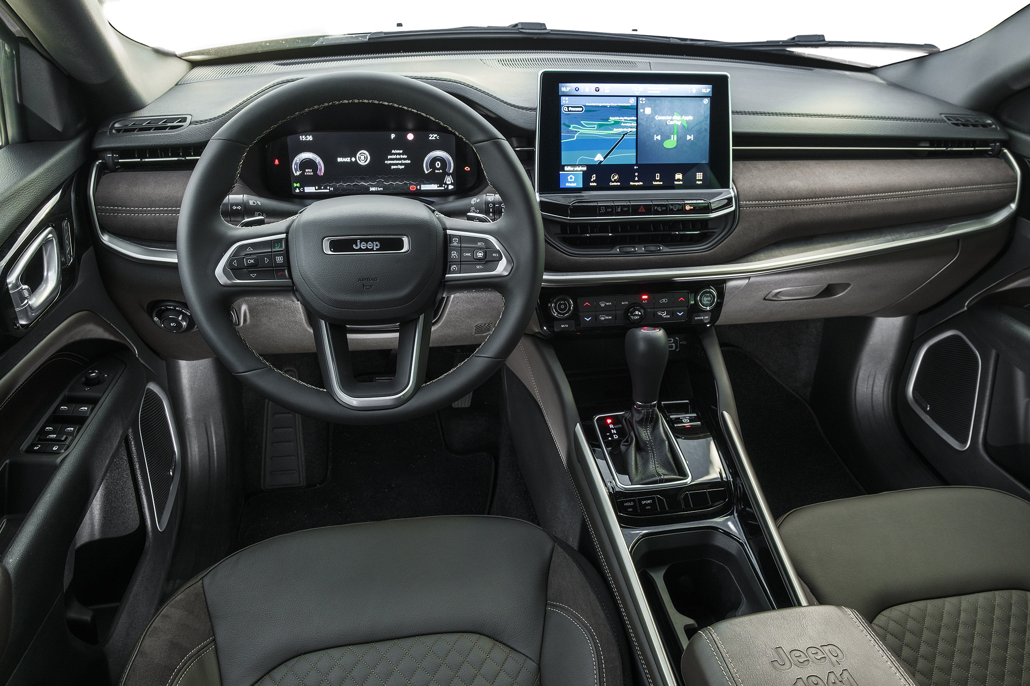 Interior de um carro Jeep, mostrando o volante multifuncional com o logo da marca, painel digital e uma tela multimídia vertical exibindo um mapa de navegação. O console central tem câmbio automático e botões de controle. Os bancos são de couro escuro com detalhes em costura.