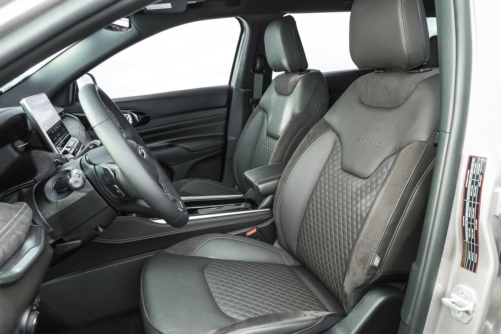 Interior de um carro com bancos de couro cinza escuro e detalhes em suede, com costuras em losango. O volante multifuncional e a tela multimídia vertical estão visíveis no painel. O banco do passageiro também é cinza escuro, com encosto de cabeça ajustável. A porta do motorista está aberta, revelando um adesivo de informações técnicas