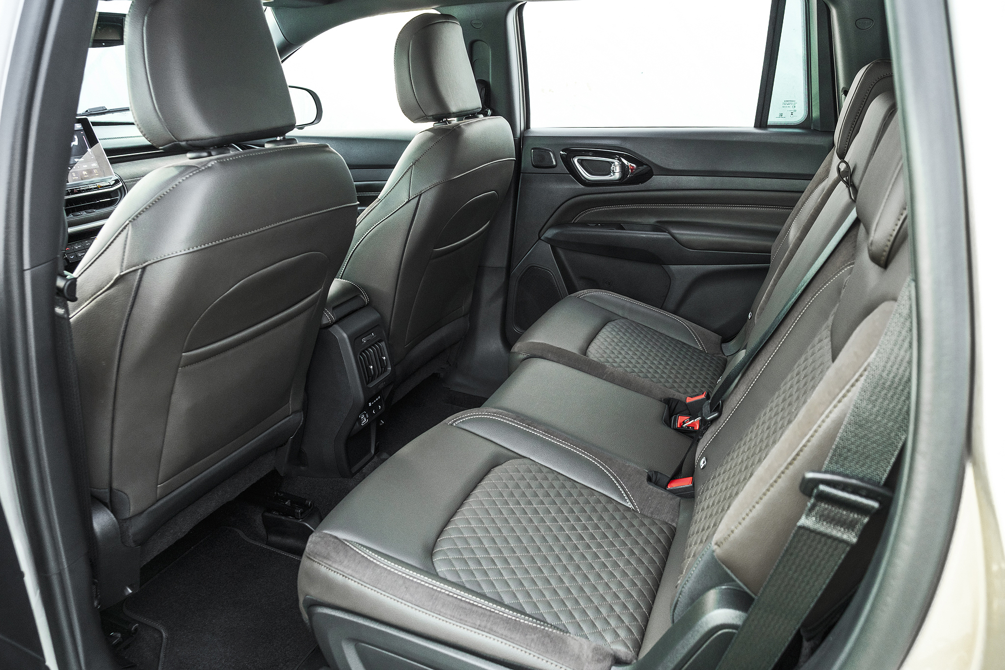 Interior de um carro com bancos de couro cinza escuro, costuras brancas e detalhes em camurça. Os bancos traseiros possuem encosto de cabeça e cintos de segurança. Ao fundo, o banco do motorista e o painel com tela digital. A porta traseira está aberta, revelando o interior espaçoso