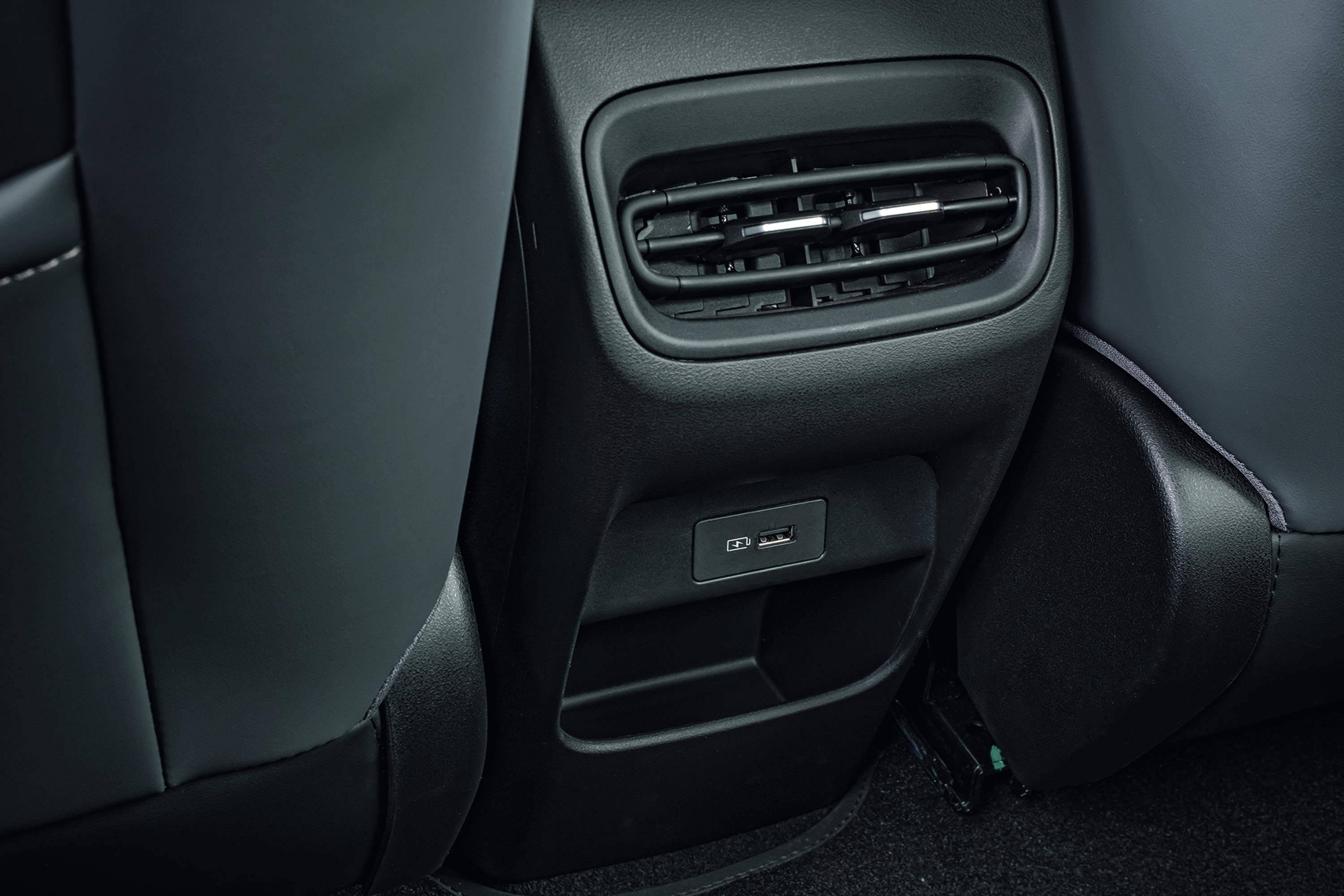 Parte traseira de um banco de carro escuro, com saídas de ar, uma porta USB-C e uma USB-A, e um porta-objetos abaixo