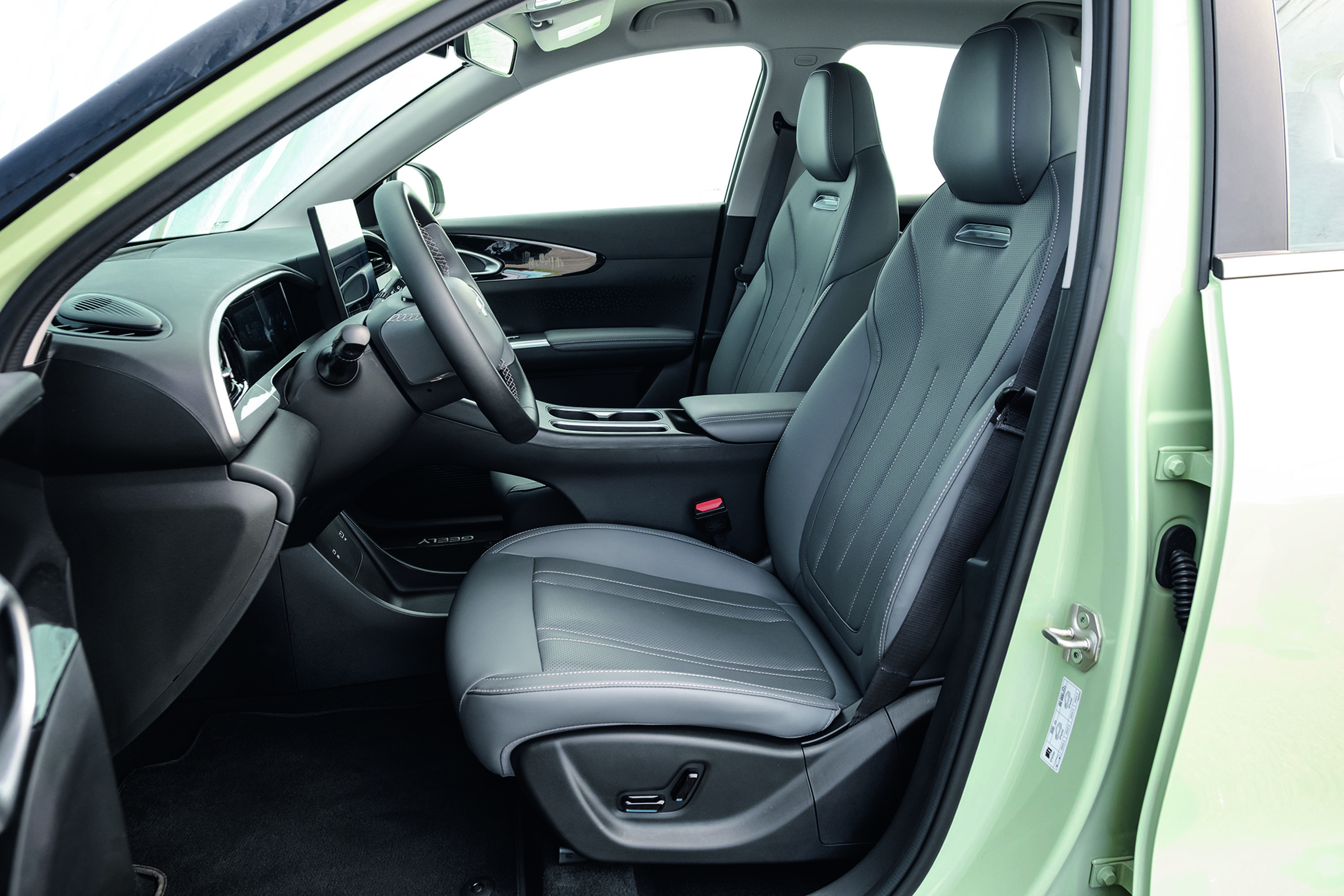 Interior de um carro verde claro, mostrando o banco do motorista e do passageiro da frente em couro cinza escuro com costuras claras. O volante preto e o painel com tela digital estão visíveis, com o chão forrado em carpete preto. A porta do motorista está aberta, revelando a pintura verde do veículo