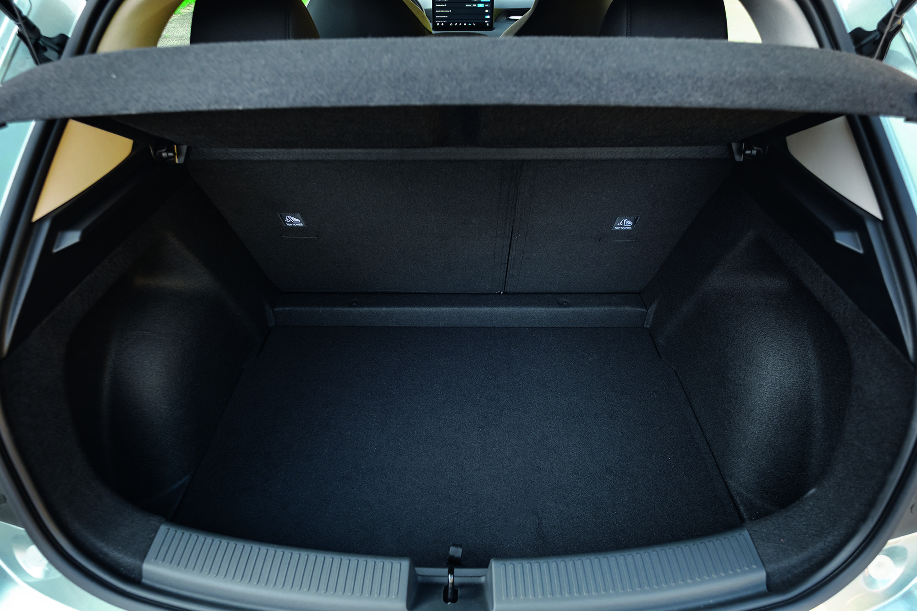 Porta-malas de um carro hatch, visto de cima, com o assoalho forrado em carpete preto e as laterais escuras. O banco traseiro está rebatido e a tampa do porta-malas está aberta, revelando o interior espaçoso do veículo. Há um pequeno detalhe bege no canto superior esquerdo e ganchos para fixação de carga.