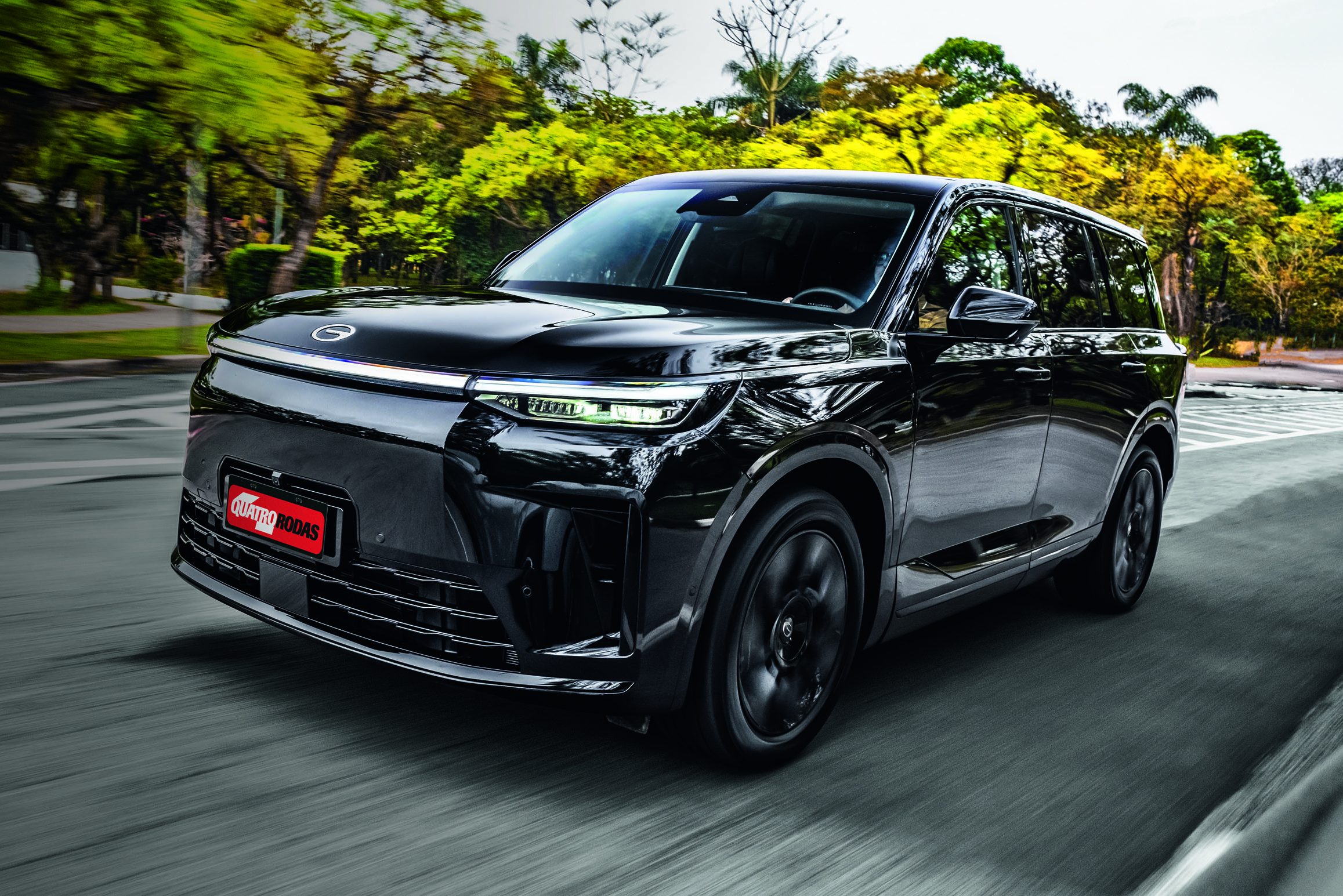 SUV preto brilhante em movimento, visto de frente e lateral, com rodas girando rapidamente. O veículo tem faróis finos e uma grade frontal fechada. Ao fundo, árvores verdes e folhagem borradas indicam velocidade