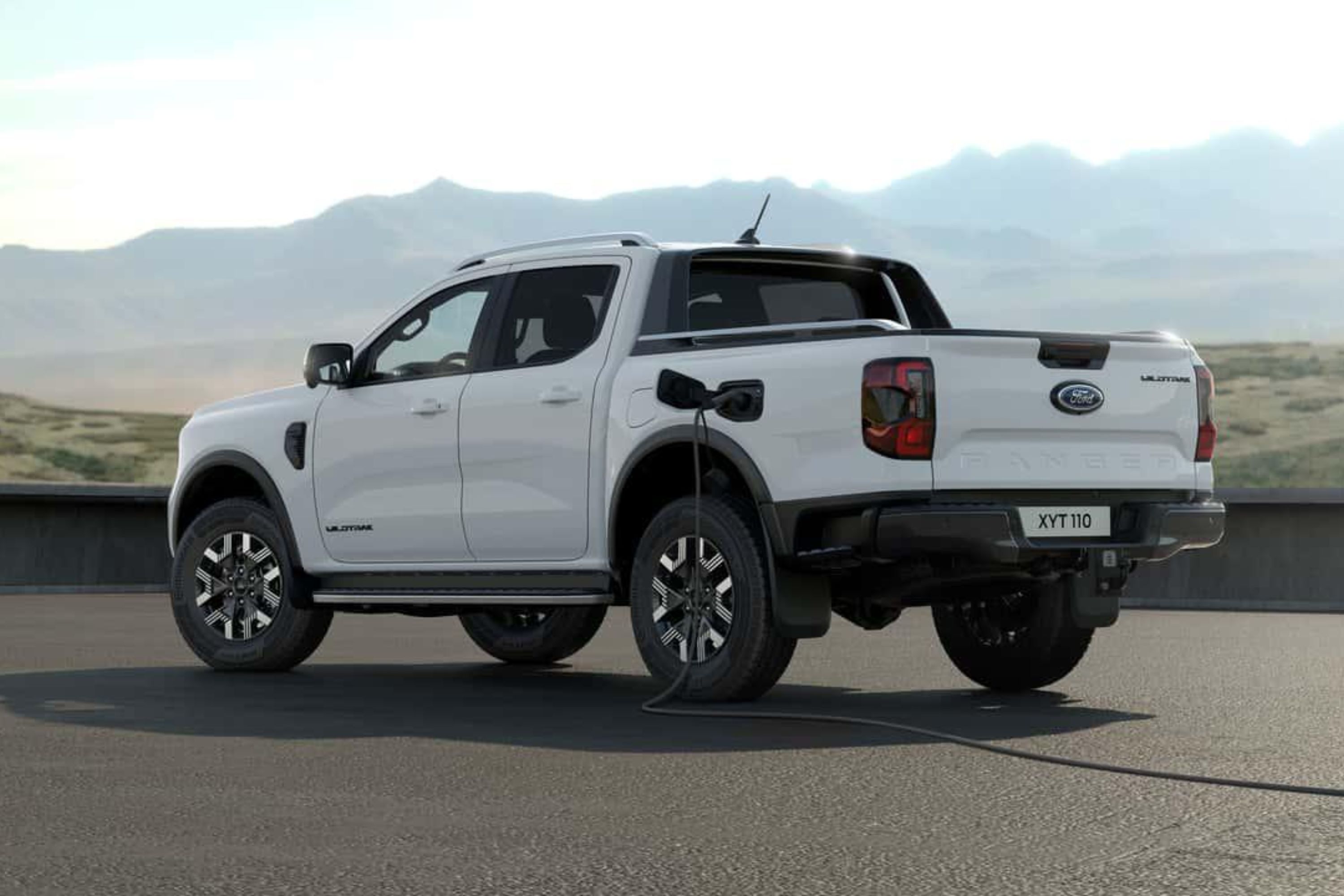 Picape branca Ford Ranger Wildtrak estacionada de costas, sendo carregada por um cabo conectado à lateral traseira, com montanhas ao fundo