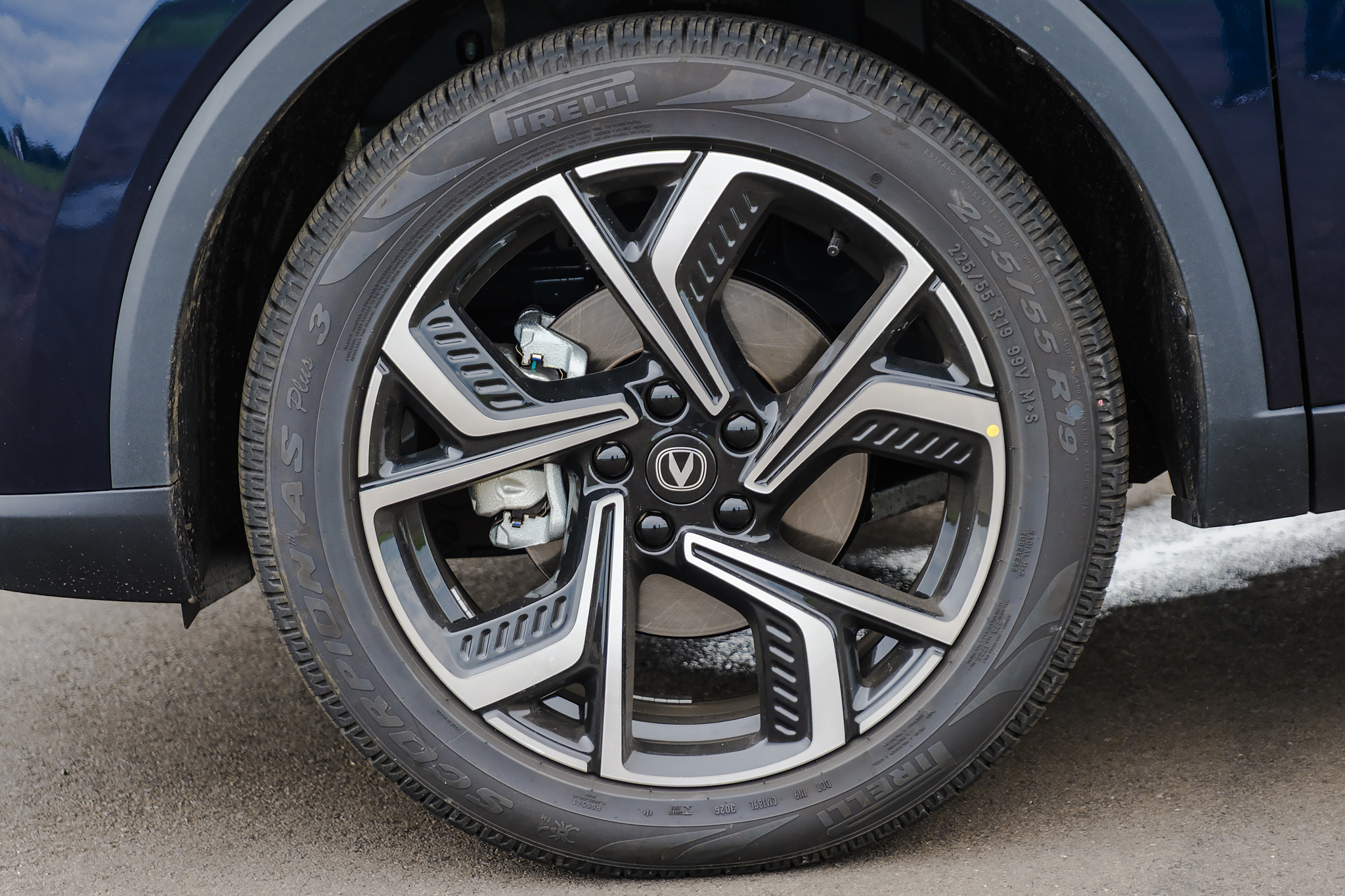 Pneu Pirelli Scorpion com aro de liga leve preto e prata, detalhe do disco de freio e pinça, parte inferior de um carro azul escuro e asfalto cinza ao fundo