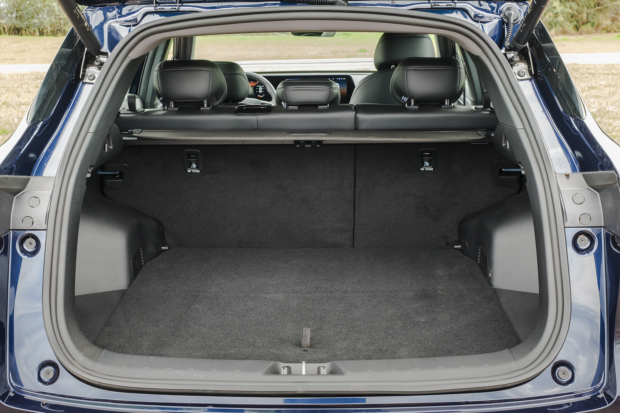 Porta-malas de um carro SUV azul escuro, visto de trás com a tampa aberta. O interior é revestido em carpete cinza escuro, com os bancos traseiros pretos visíveis ao fundo, rebatidos para aumentar o espaço de carga. Há ganchos de fixação nas laterais e uma barra de cobertura retrátil acima dos bancos