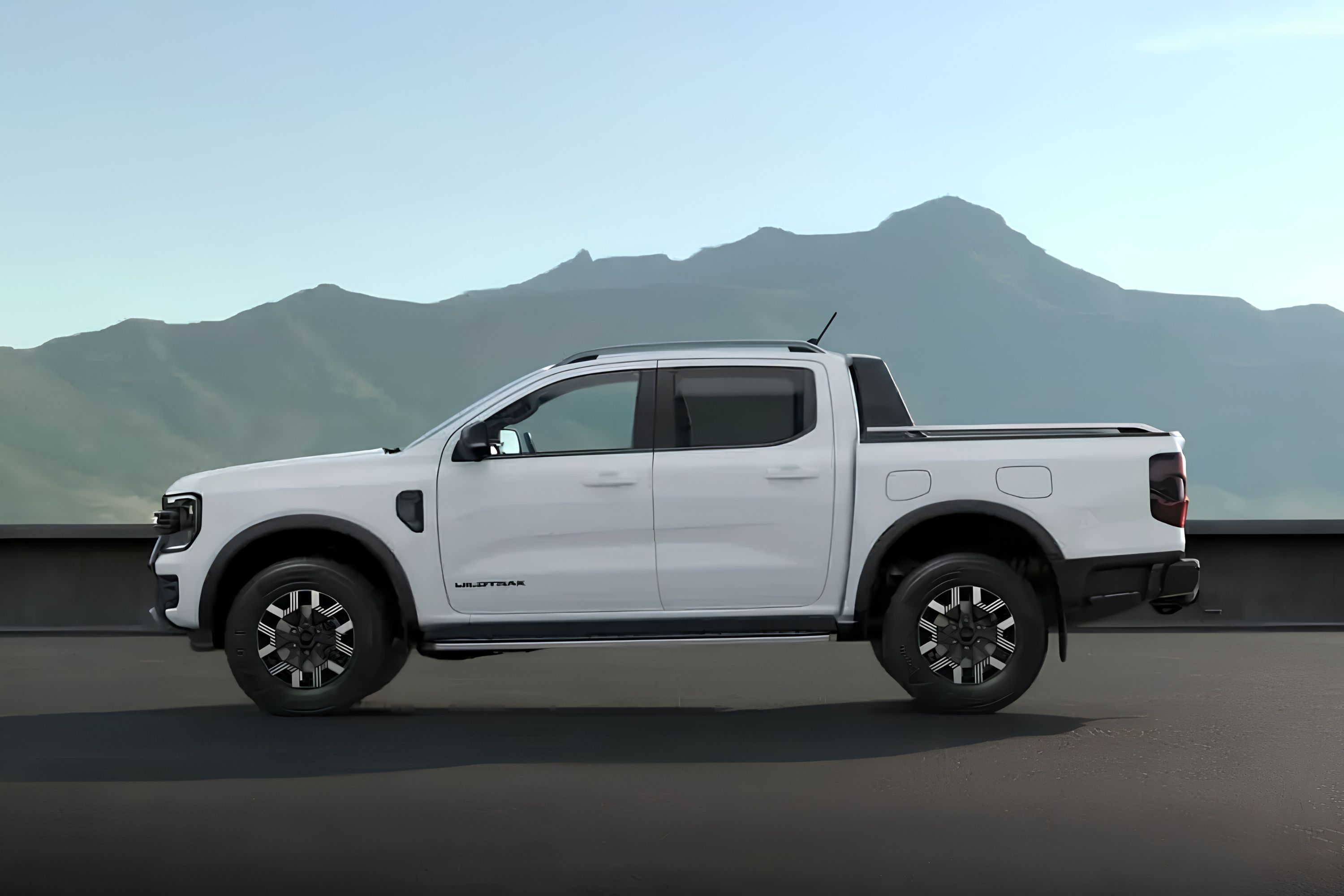 Picape branca de quatro portas, modelo Wildtrak, vista de perfil, estacionada em uma superfície escura com montanhas ao fundo sob um céu claro