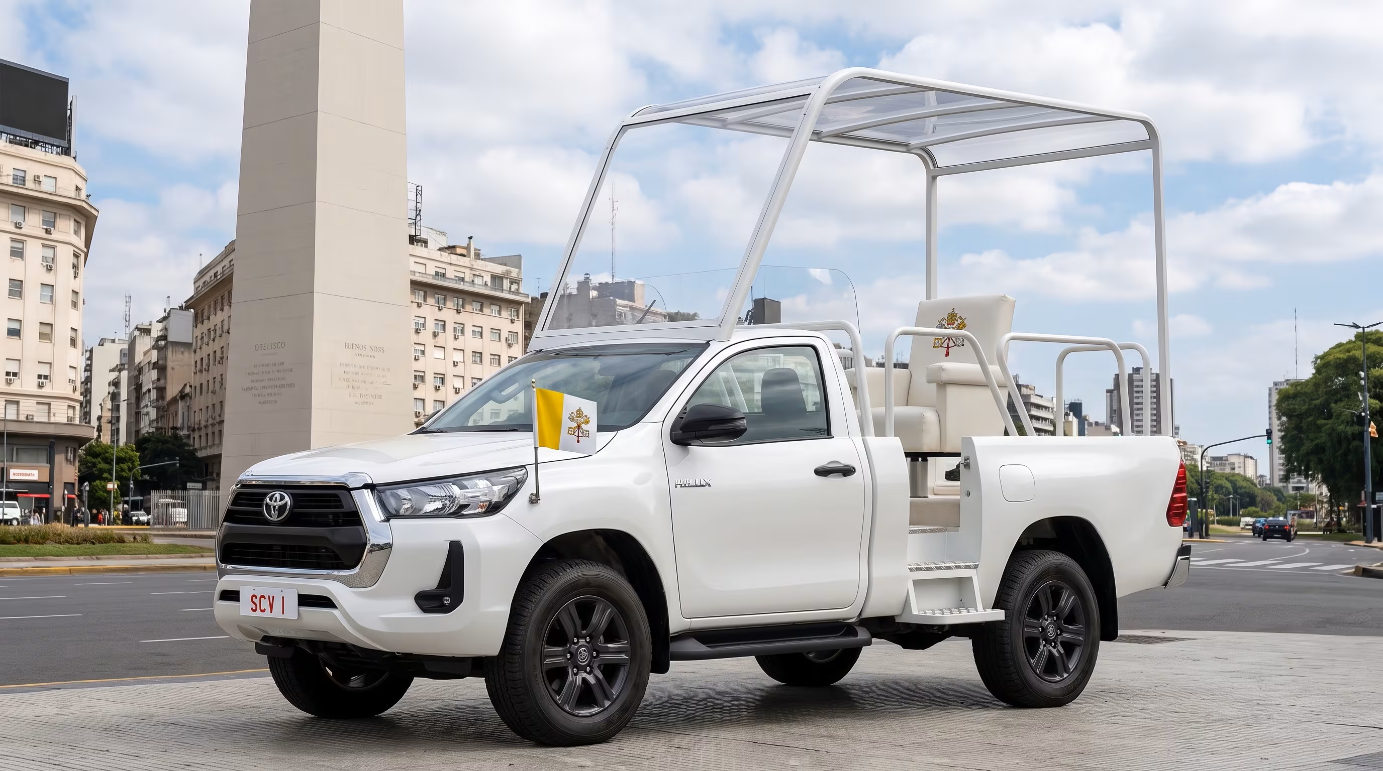 Papamóvel branco, uma picape Toyota Hilux adaptada com assento elevado e cobertura transparente, estacionado em uma rua urbana. Uma bandeira do Vaticano está na frente, e o Obelisco de Buenos Aires e prédios históricos aparecem ao fundo sob céu azul com nuvens