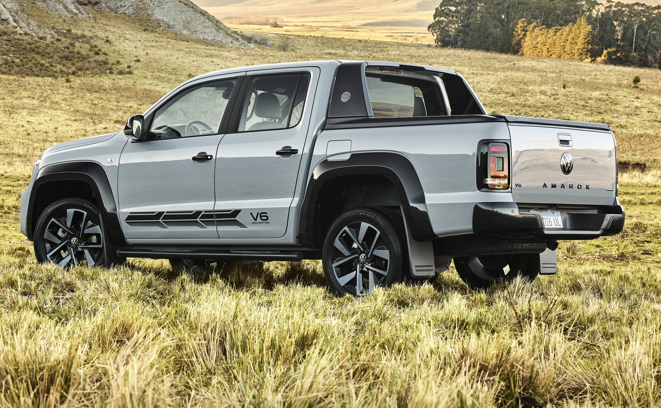 Picape Volkswagen Amarok cinza-claro, com detalhes pretos e adesivo V6 na lateral, estacionada em um campo de grama seca, com colinas e árvores ao fundo