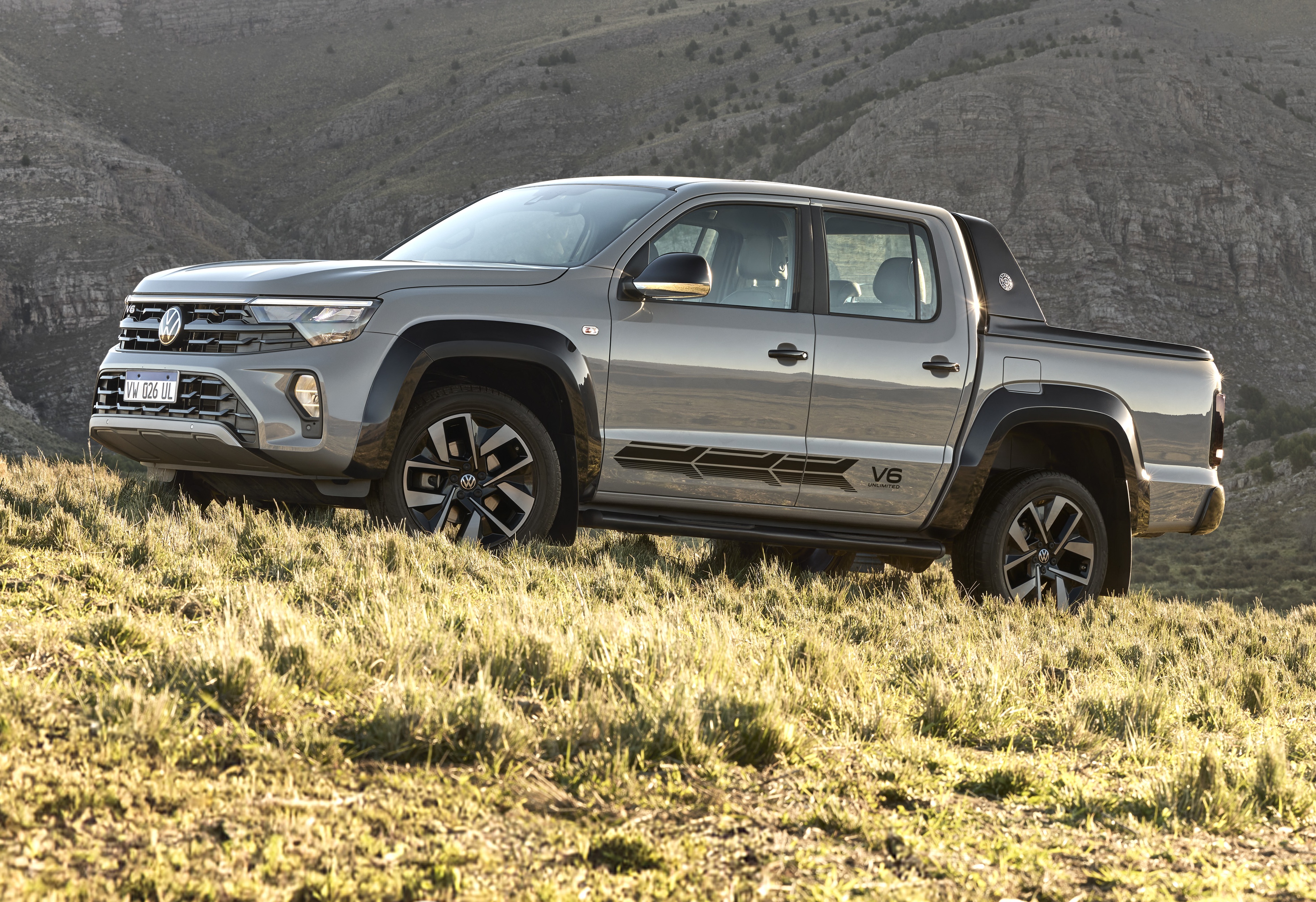 Picape Volkswagen Amarok cinza-chumbo estacionada em uma colina de grama seca, com montanhas rochosas ao fundo. O veículo tem detalhes em preto, incluindo a grade frontal, para-lamas e um adesivo lateral com V6 e listras geométricas