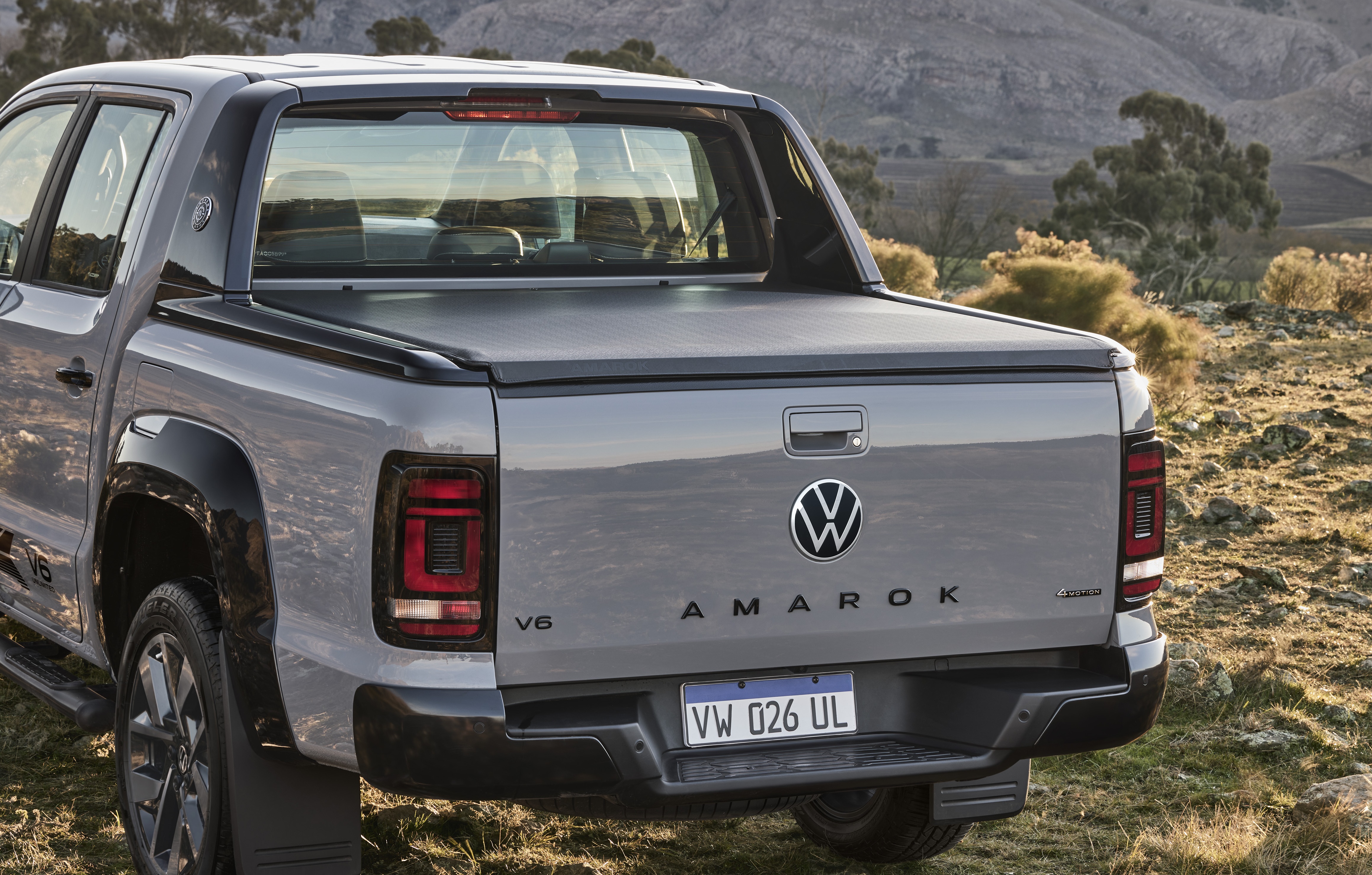 Traseira de uma picape Volkswagen Amarok cinza, com capota marítima preta, estacionada em terreno árido com vegetação rasteira e montanhas ao fundo. A placa é VW 026 UL