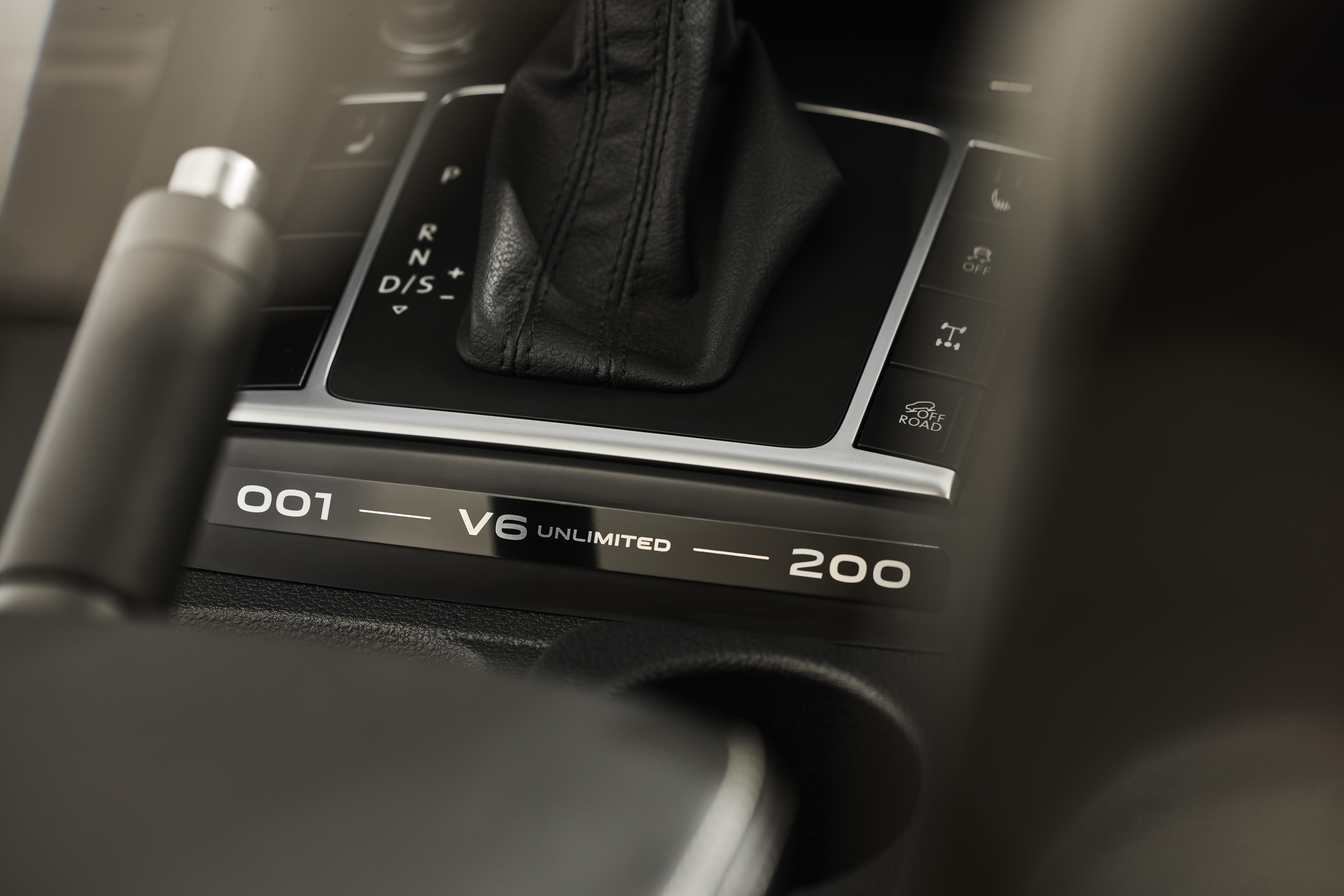 Detalhe do console central de um carro, com a alavanca de câmbio automático em couro preto, freio de mão e botões de controle. Uma placa metálica exibe 001 — V6 UNLIMITED — 200