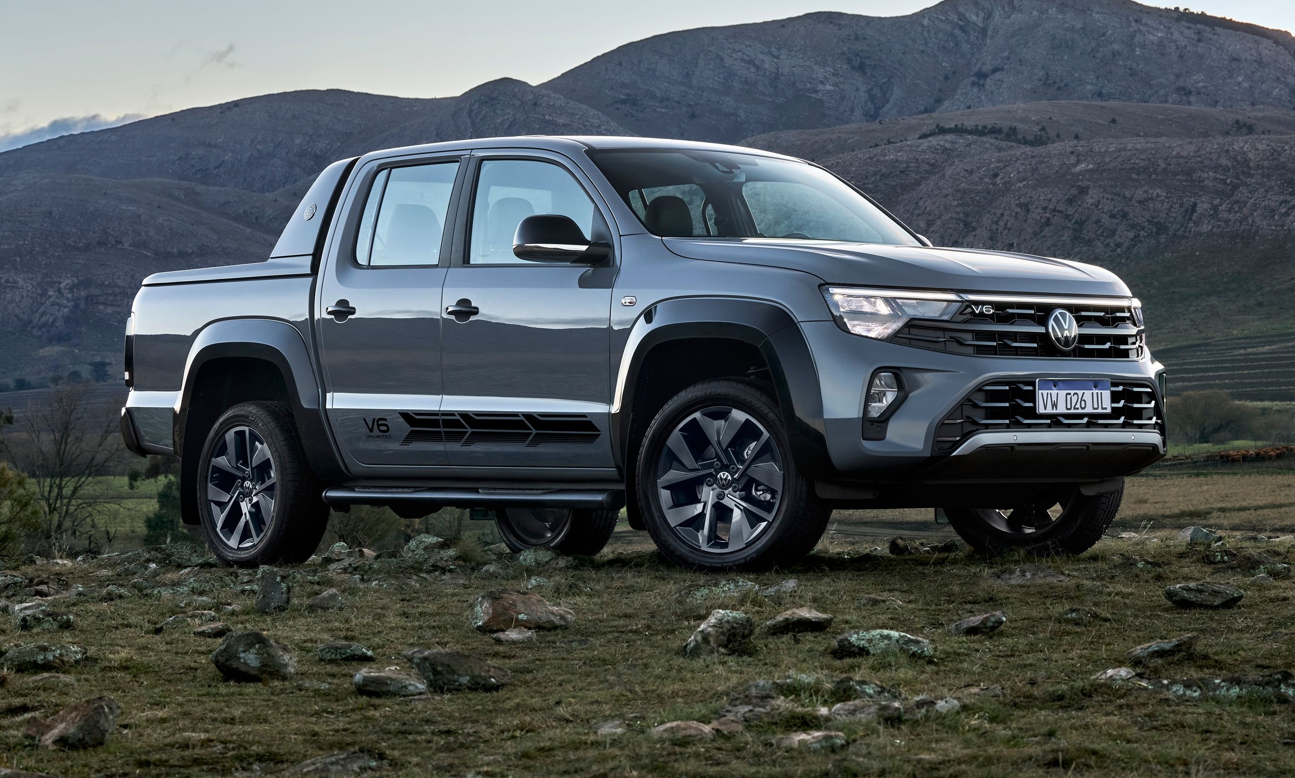 Picape Volkswagen Amarok cinza-escura, modelo V6, estacionada em terreno rochoso com vegetação rasteira, montanhas ao fundo sob céu claro