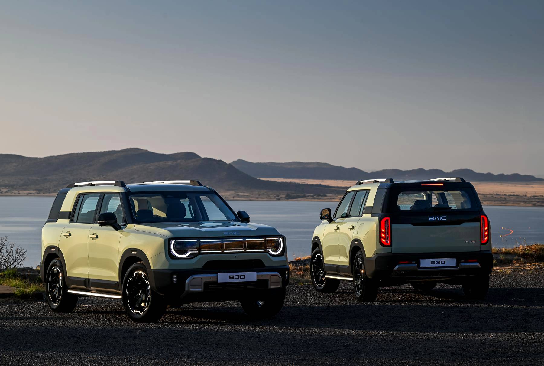 Dois SUVs BAIC B80, um de frente e outro de costas, em tom verde-claro, estacionados em um terreno escuro com um lago e montanhas ao fundo sob um céu claro