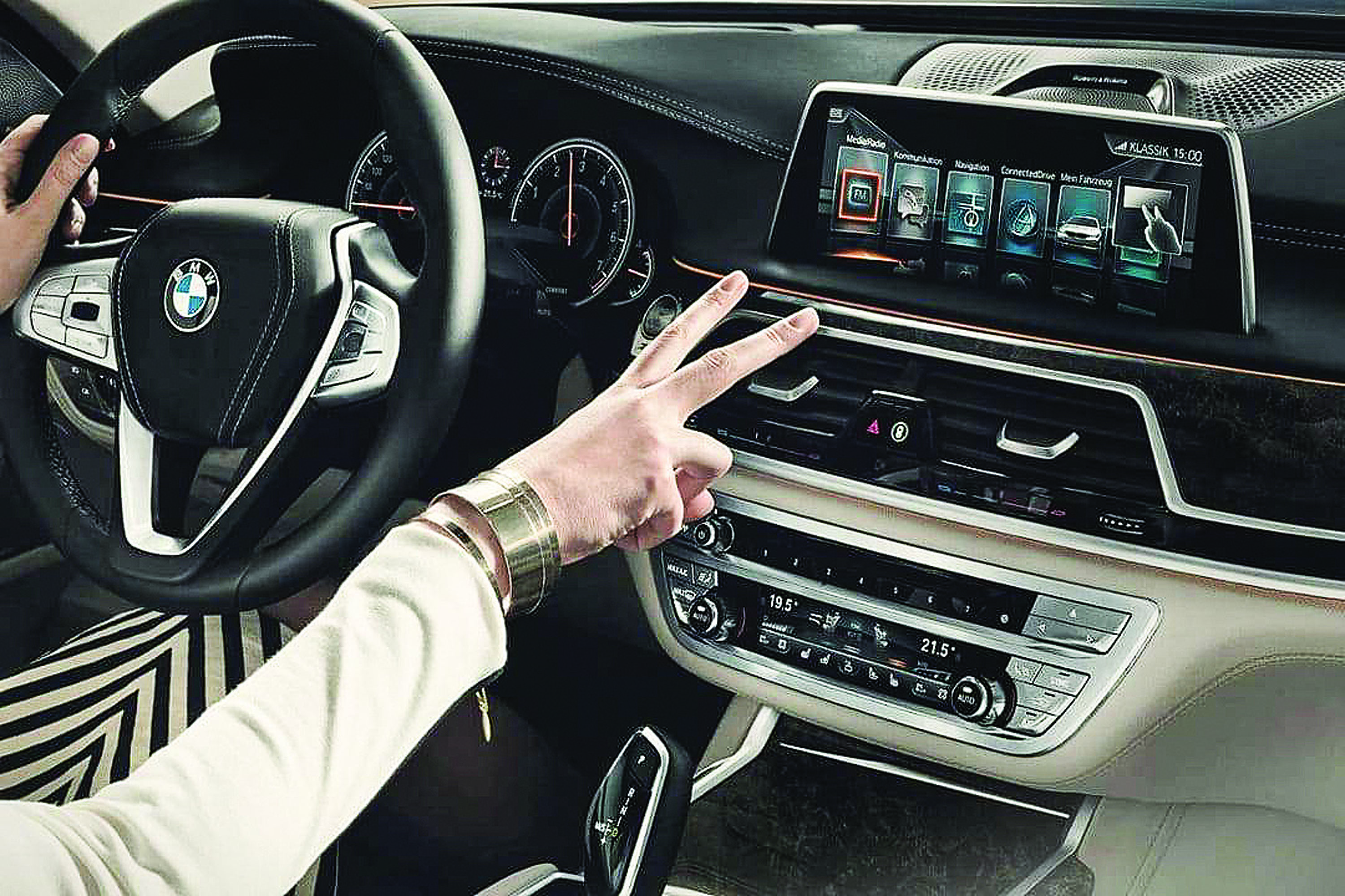 Mãos de uma pessoa dirigindo um carro de luxo, com uma mão no volante e a outra fazendo um gesto de paz e amor em frente ao painel digital e controles de ar condicionado