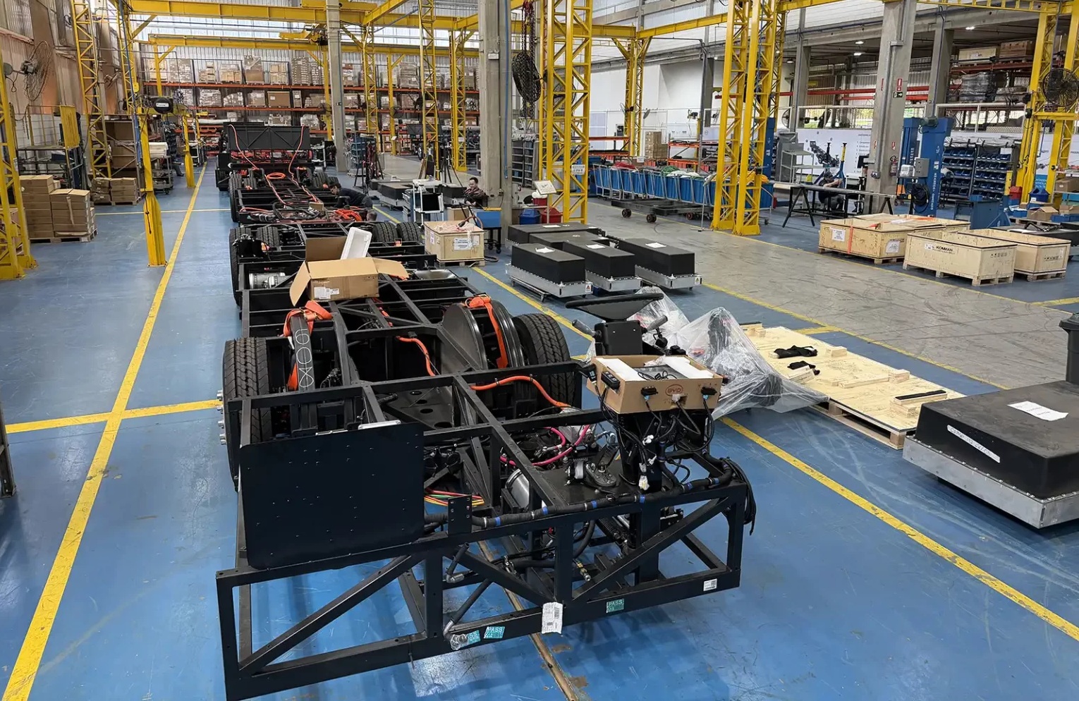 Chassis de ônibus elétricos em linha de montagem industrial, com estruturas metálicas pretas, pneus e fiação laranja. Caixas de papelão e madeira, além de componentes embalados, estão dispostos no chão azul com faixas amarelas, sob uma estrutura metálica amarela no teto