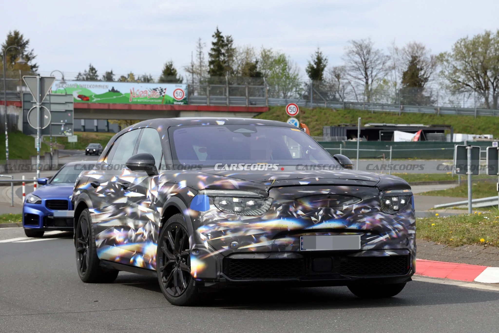 Um carro SUV camuflado com padrão de diamantes coloridos e rodas pretas, em uma pista de testes. Ao fundo, um carro azul e uma ponte com outdoor verde. O céu está nublado