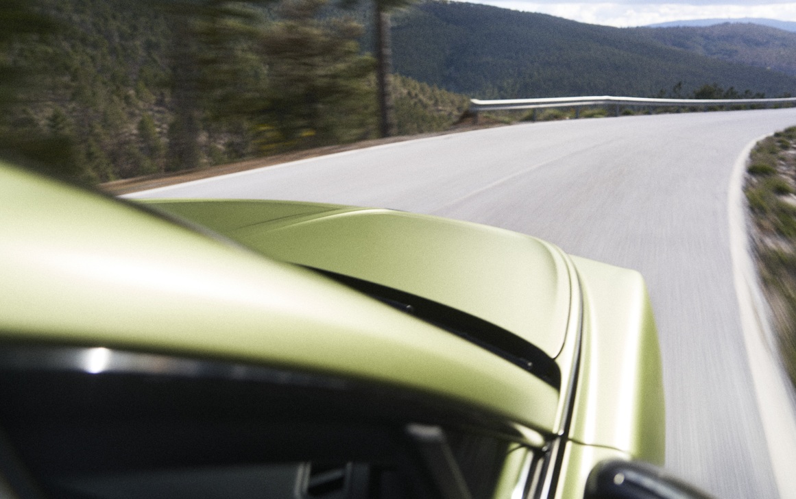 Vista superior de um carro verde-limão em movimento, virando à direita em uma estrada sinuosa com montanhas arborizadas ao fundo e céu nublado