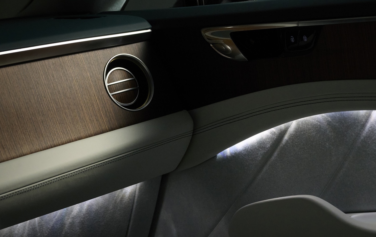 Interior de carro luxuoso com painel de madeira escura, saída de ar circular cromada e estofamento claro com costura aparente. Uma faixa de luz ambiente branca ilumina a parte inferior da porta e o assento, criando um contraste elegante