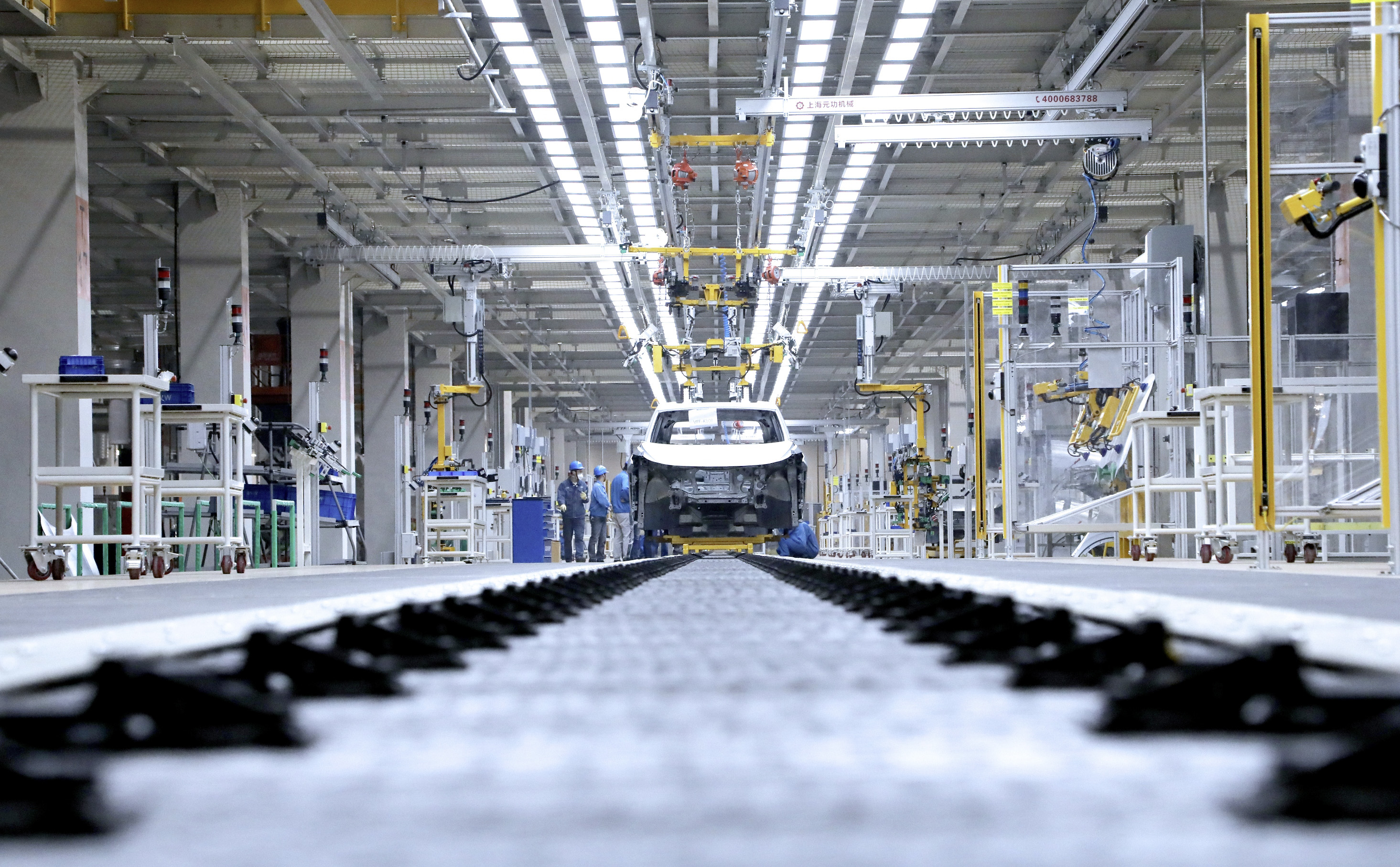 Linha de montagem de carros com um chassi branco no centro, cercado por trabalhadores de capacete azul e máquinas industriais. O chão tem uma esteira transportadora em primeiro plano, e o teto é alto com iluminação e estruturas metálicas.