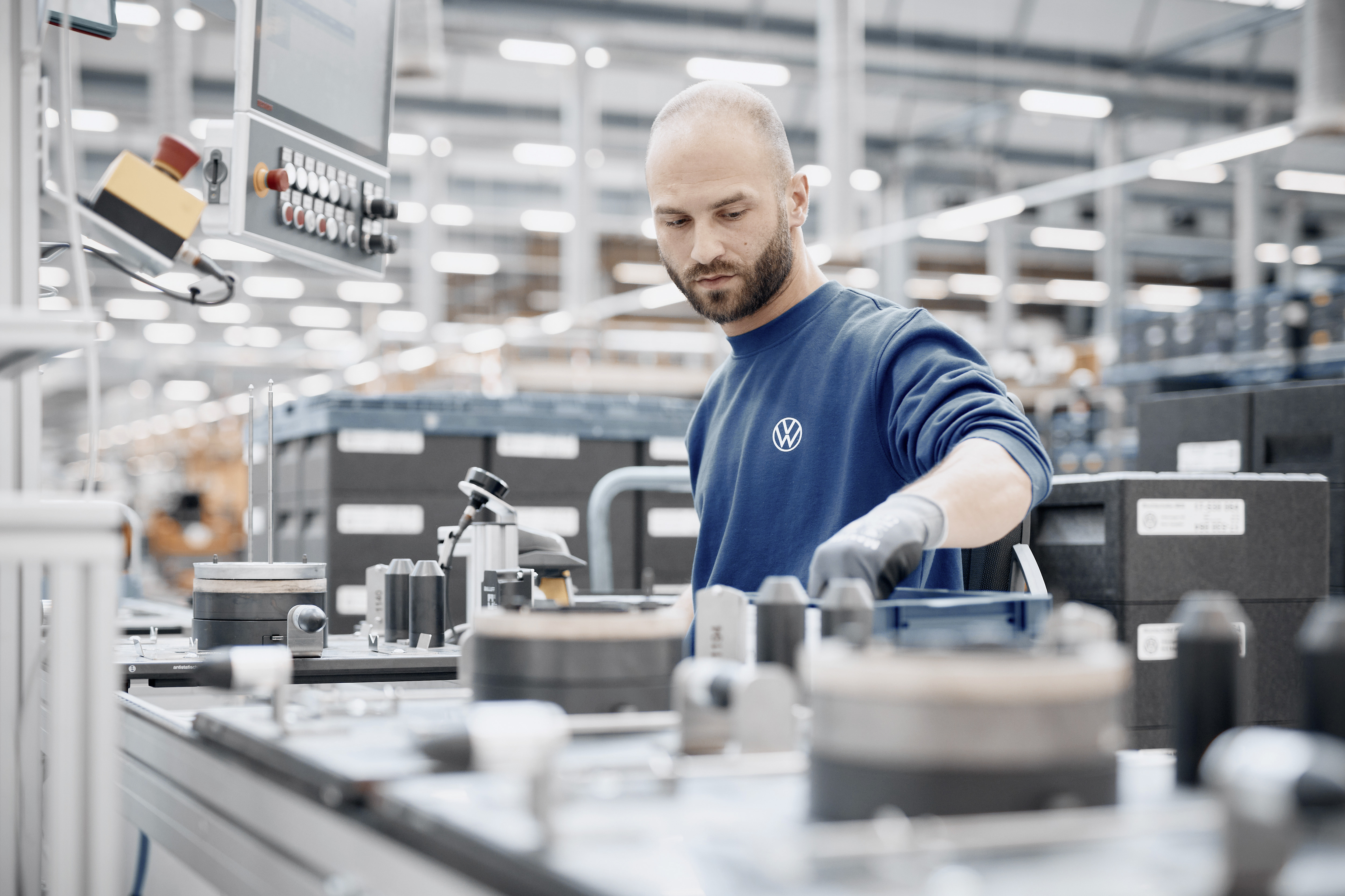 Homem barbudo, de suéter azul com logo da Volkswagen e luvas cinzas, opera equipamentos em uma linha de montagem industrial
