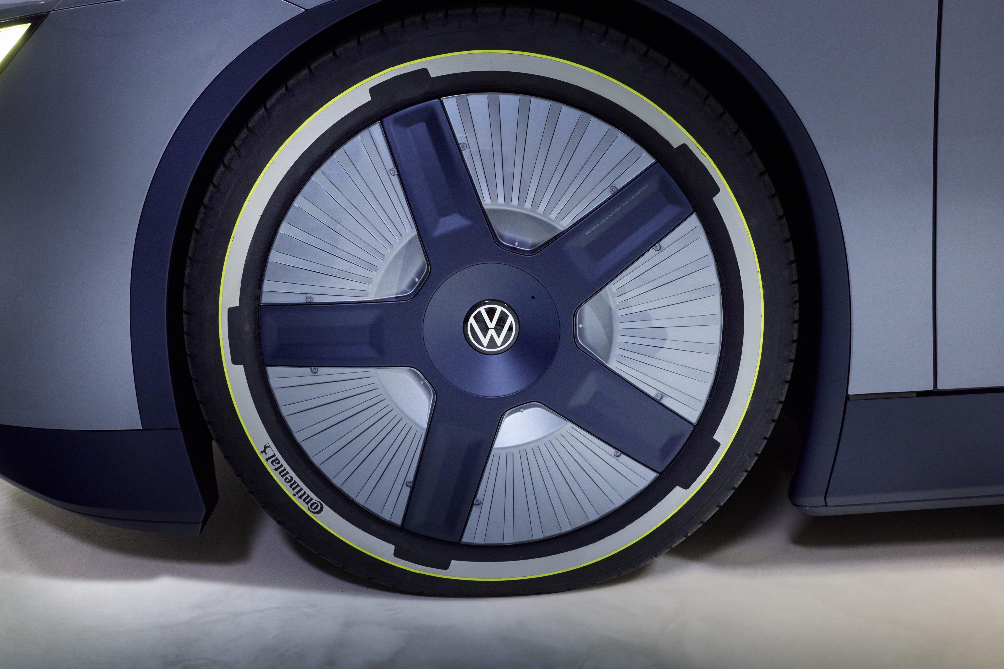 Roda de carro cinza-escuro com calota azul-marinho de cinco raios e logo Volkswagen, pneu Continental com faixa amarela e cinza, parte de um veículo moderno