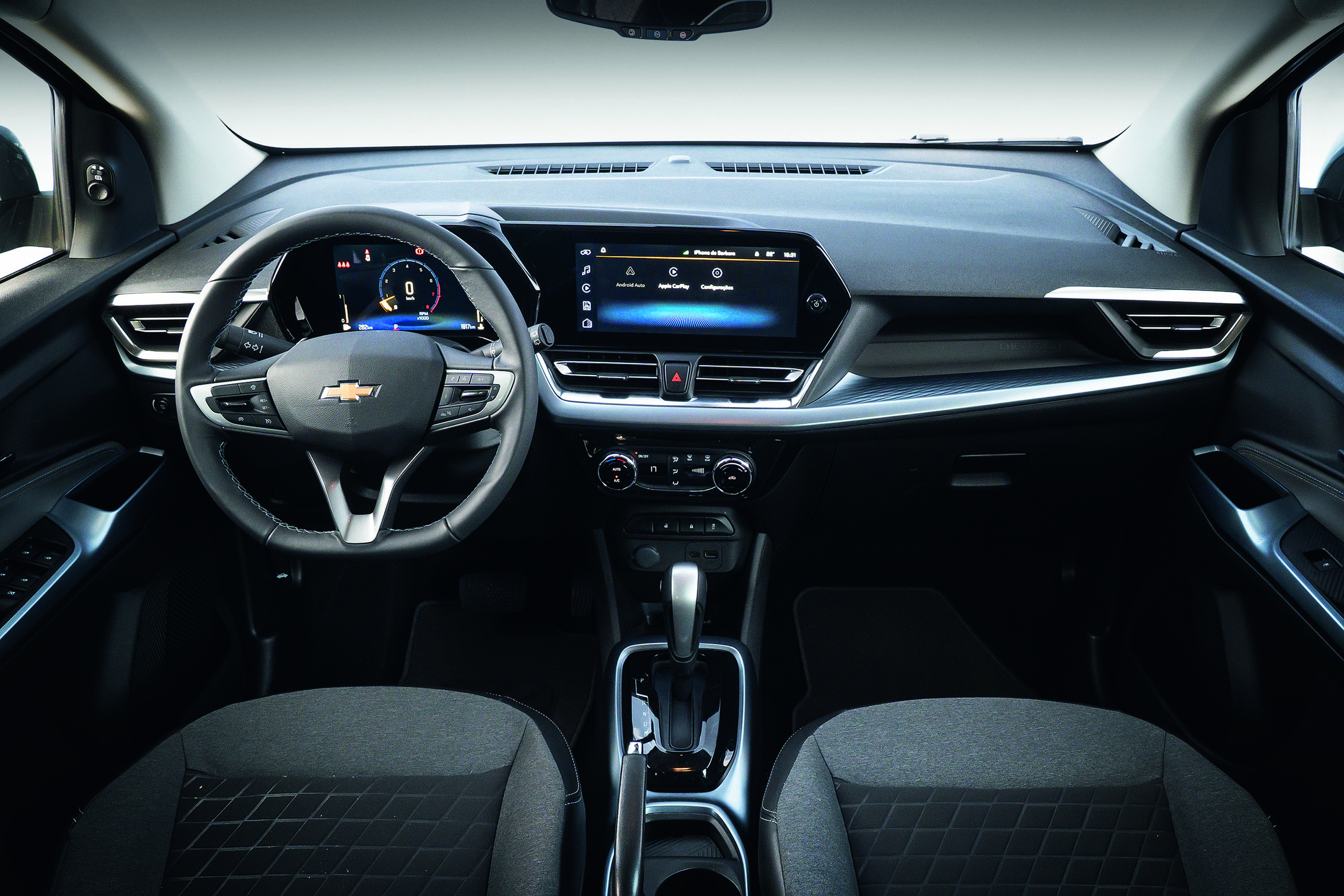 Interior de um carro moderno, com volante multifuncional da Chevrolet à esquerda e painel digital. A central multimídia exibe um menu com ícones e um fundo azul. Detalhes em prata e preto compõem o design do painel, com saídas de ar e controles de climatização. Os bancos dianteiros, cinza escuro com padrão geométrico, são visíveis em primeiro plano. O câmbio automático está no console central