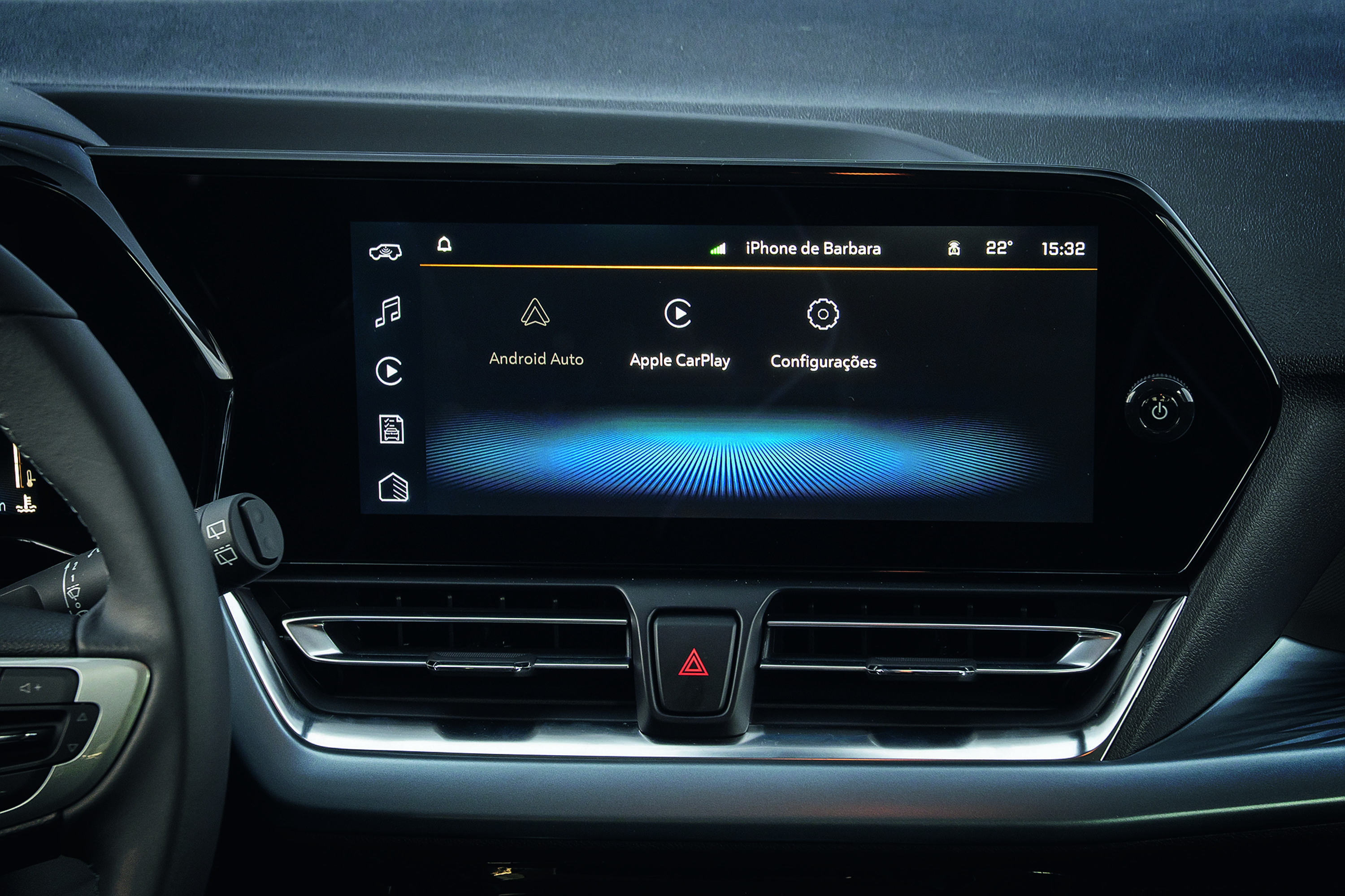 Tela multimídia de carro com fundo escuro, exibindo ícones de aplicativos como Android Auto, Apple CarPlay e Configurações. No canto superior direito, lê-se iPhone de Barbara, 22° e 15:32. Abaixo da tela, saídas de ar e um botão de alerta vermelho. À esquerda, parte do volante e alavanca de seta.