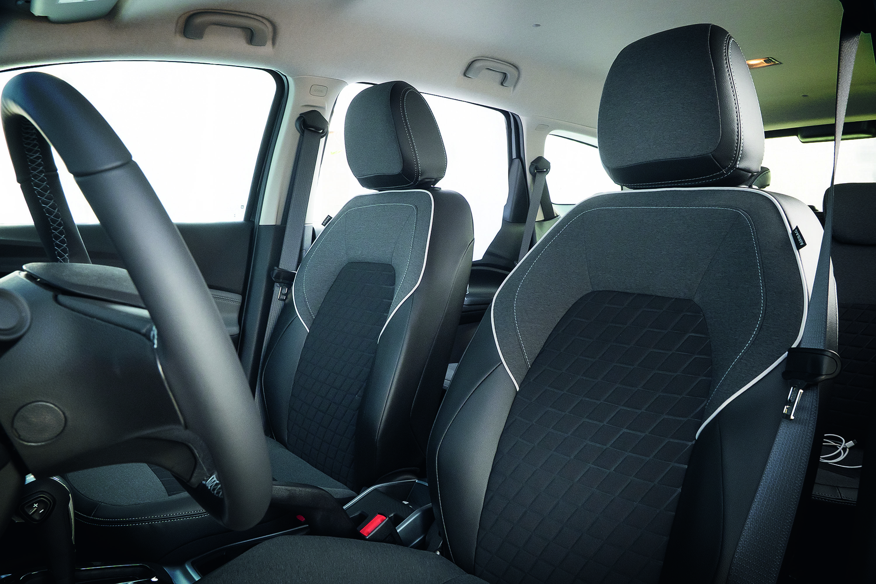 Interior de carro com volante preto à esquerda e dois bancos dianteiros cinza escuro com detalhes em branco e textura geométrica, cintos de segurança visíveis