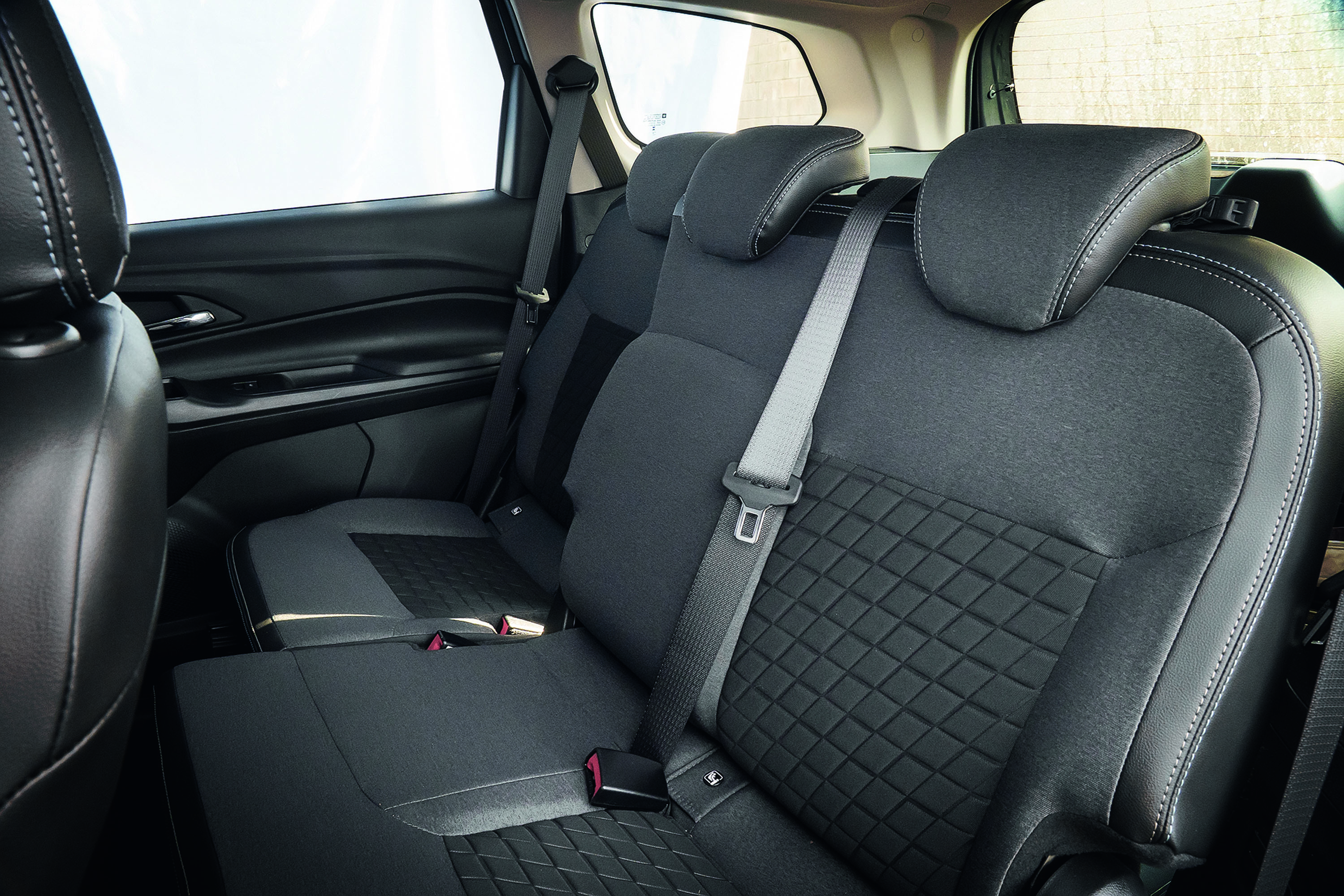 Interior de carro com bancos traseiros em tecido cinza escuro e detalhes em couro preto, com costuras brancas. O banco do meio tem cinto de segurança cinza claro afivelado. A janela lateral está visível ao fundo