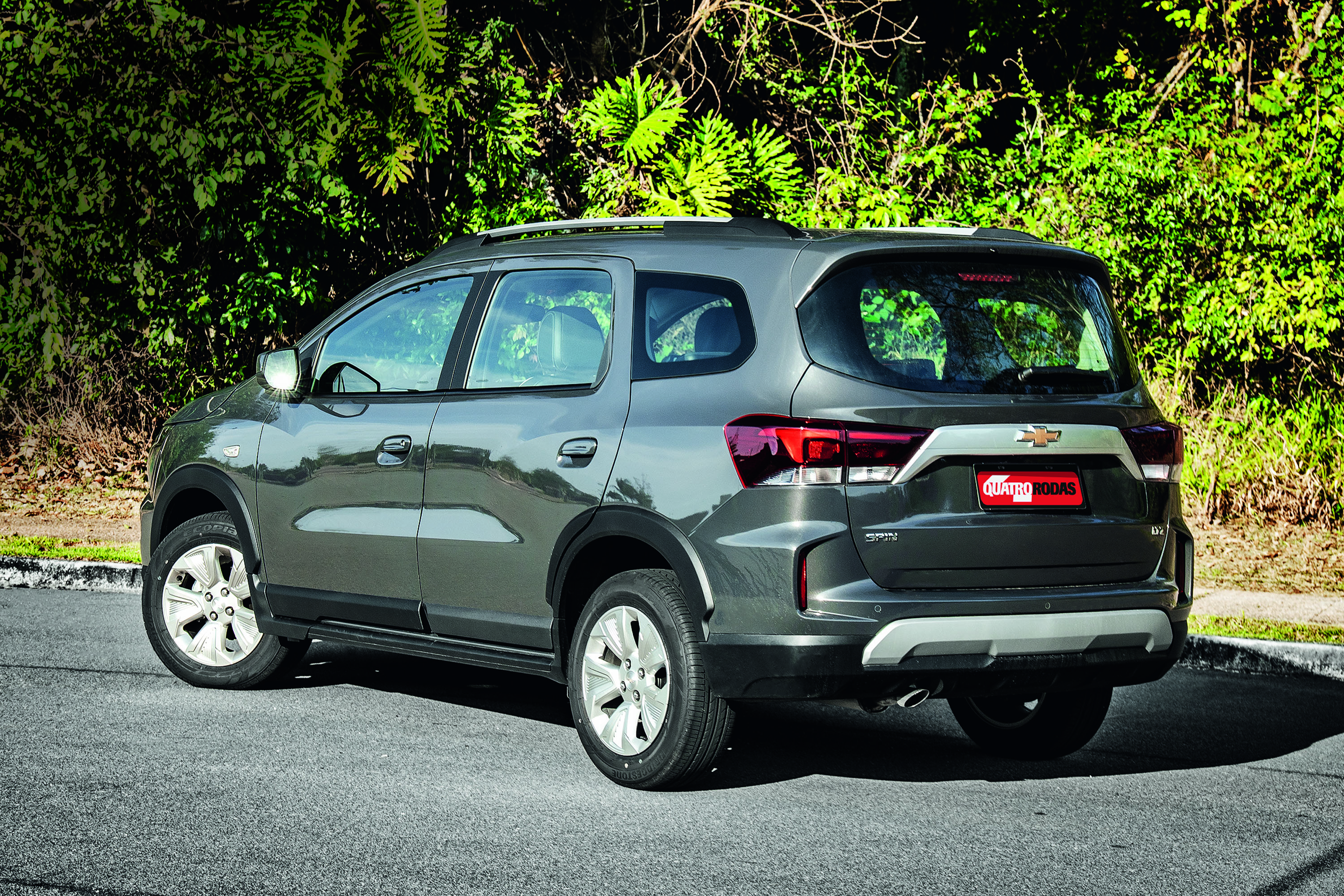 SUV cinza-escuro, modelo Chevrolet Spin, estacionado de lado em uma rua asfaltada, com vegetação verde ao fundo. O carro tem rack de teto prateado, rodas de liga leve e placa vermelha QUATRO RODAS