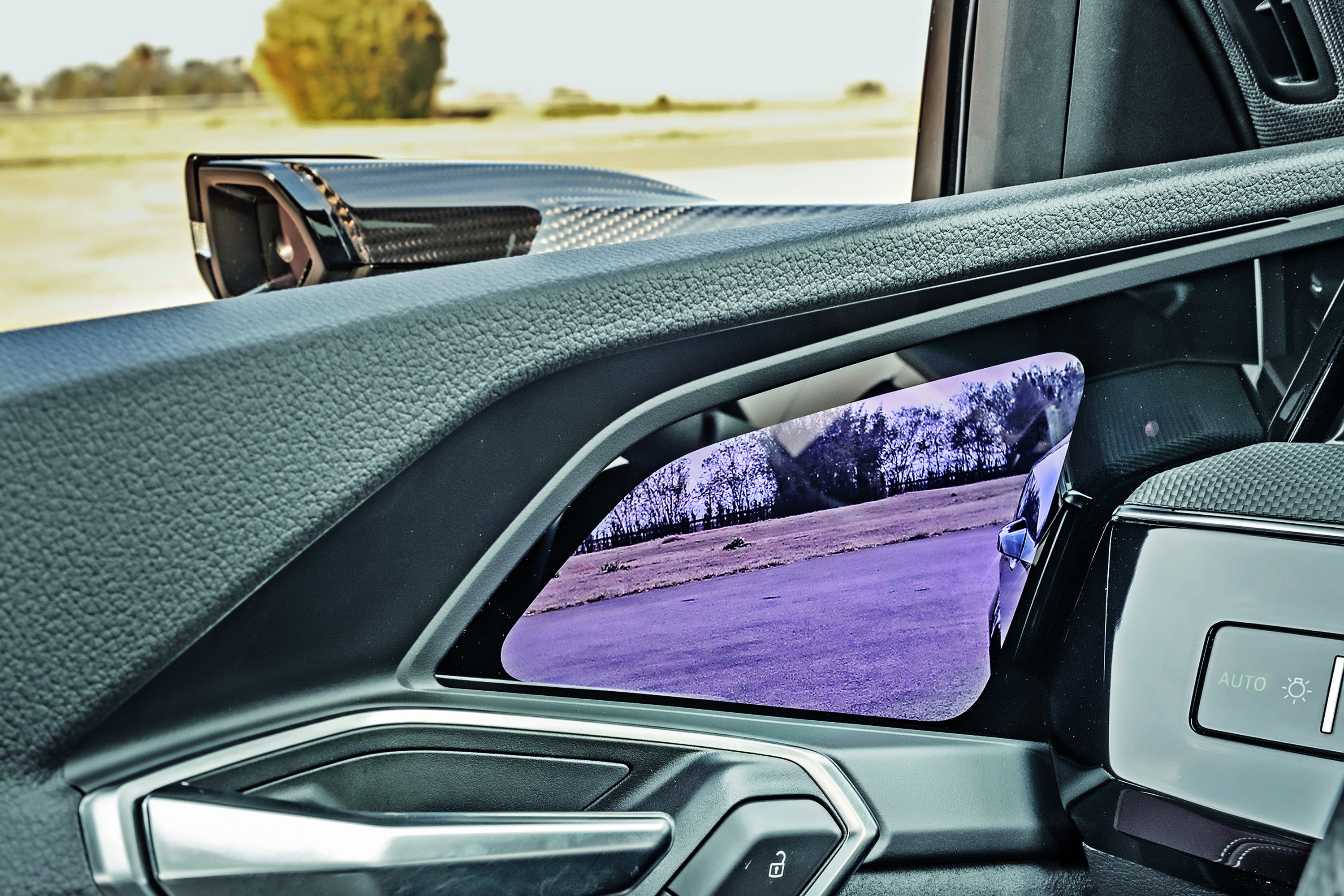 Detalhe do interior de um carro, mostrando o espelho retrovisor digital que reflete uma paisagem árida com árvores e um campo roxo. A maçaneta da porta e os controles do vidro estão visíveis