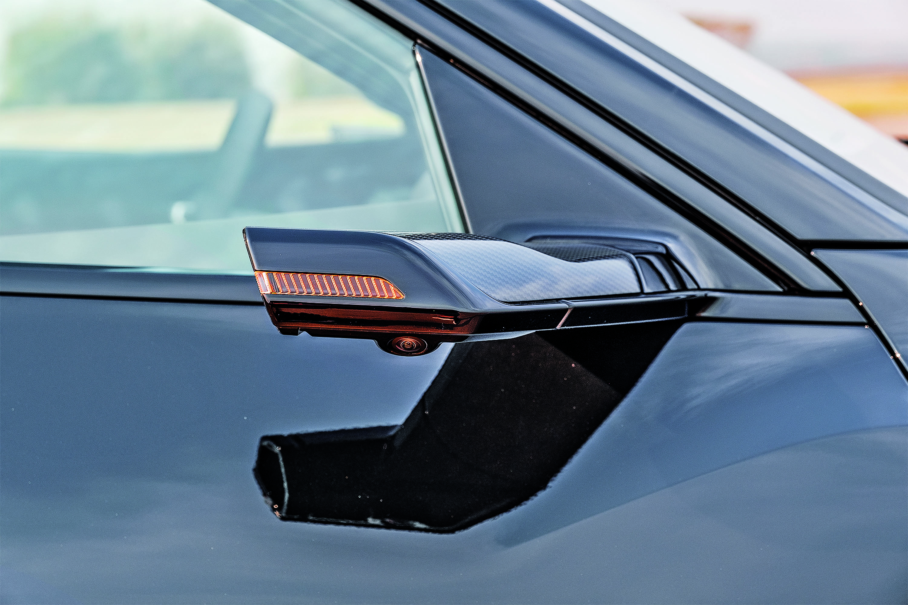 Retrovisor lateral de um carro preto brilhante, com luzes de seta laranja acesas e uma pequena câmera na parte inferior. O para-brisa e a janela lateral refletem o céu claro e a paisagem desfocada