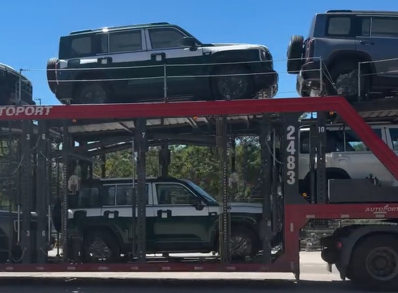 Um caminhão-cegonha vermelho transporta vários veículos utilitários esportivos, incluindo um preto e branco no nível superior e outro preto e branco no nível inferior. O caminhão exibe o número 2483 na lateral e a palavra AUTOPORT em vermelho. O céu é azul claro e há árvores ao fundo