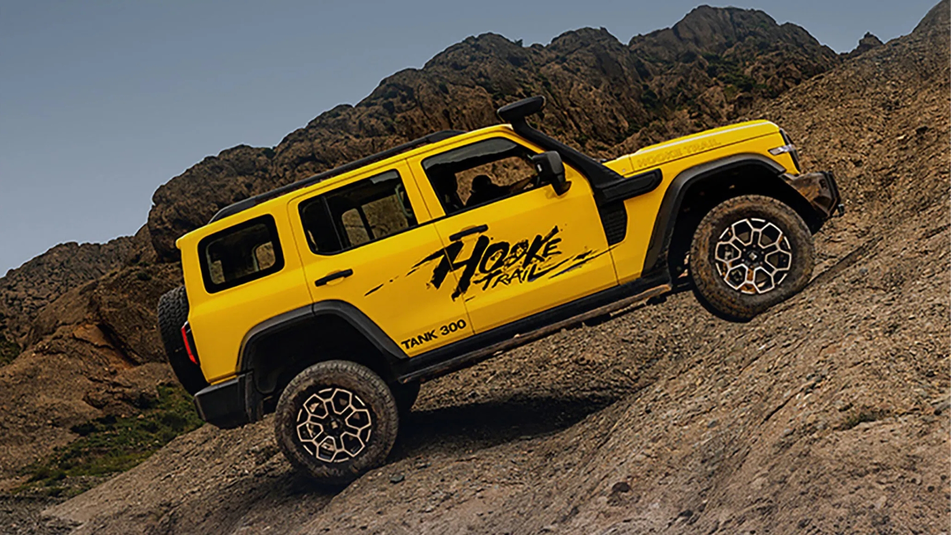 Jipe amarelo com adesivo Hooke Trail e Tank 300 na lateral, subindo uma encosta rochosa íngreme, com montanhas ao fundo e céu azul