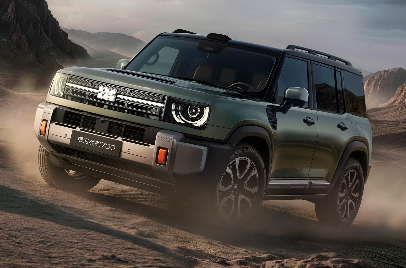 SUV verde-escuro com teto preto e rack de teto, subindo uma trilha de terra em um cenário desértico com montanhas ao fundo. A placa frontal tem caracteres chineses e o número 700. Poeira levanta sob os pneus.