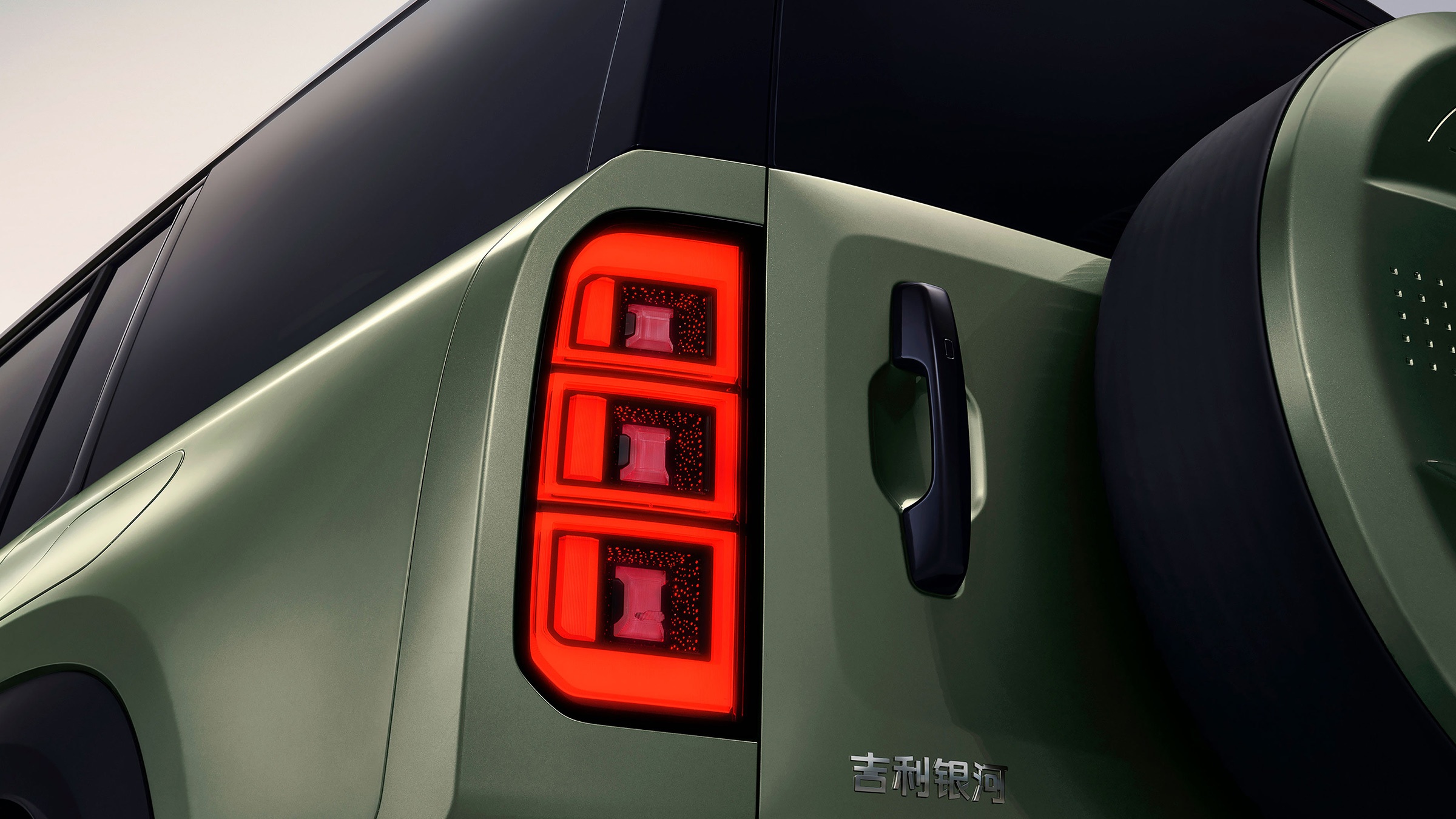 Traseira de um carro verde-oliva com lanternas LED verticais acesas em vermelho, divididas em três blocos retangulares. À direita, maçaneta preta e parte do estepe coberto, com o nome 吉利银河 em chinês na lataria