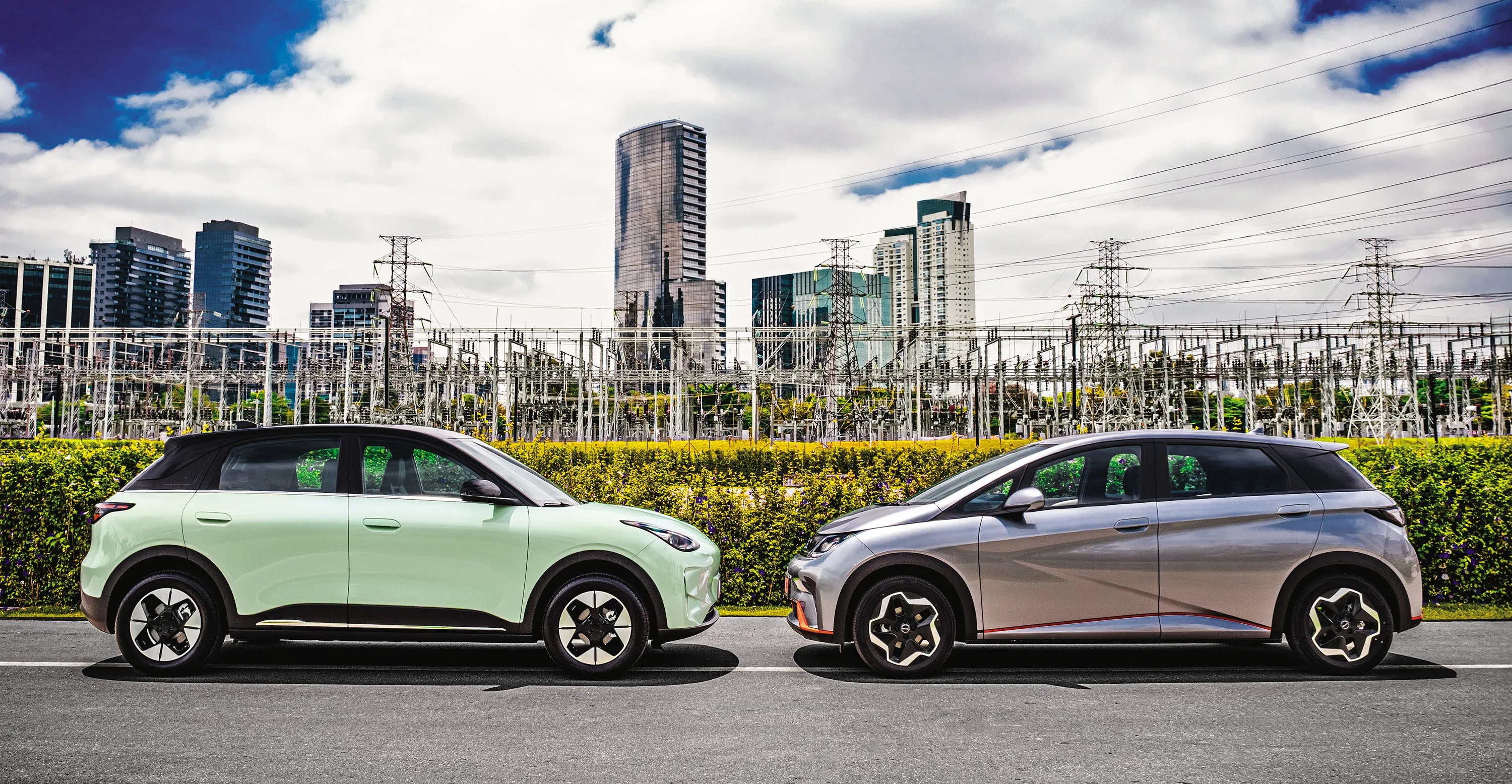 Dois carros elétricos, um verde-claro e outro prata, estacionados frente a frente em uma rua. Ao fundo, uma subestação de energia com torres e fios, e prédios altos sob um céu parcialmente nublado