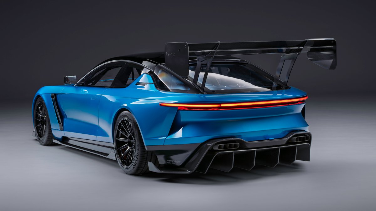 Carro esportivo azul-claro com teto preto, visto de trás e lateralmente, exibindo um grande aerofólio traseiro preto, lanterna horizontal vermelha e difusor aerodinâmico na parte inferior. As rodas são pretas com múltiplos raios, e o fundo é cinza escuro