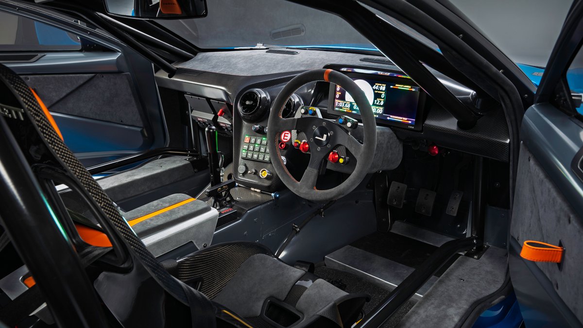 Interior de um carro de corrida azul, com bancos esportivos pretos e detalhes laranja. O volante é cinza escuro com botões coloridos, e o painel possui uma tela digital exibindo informações. O habitáculo tem estrutura de segurança e pedais de metal.