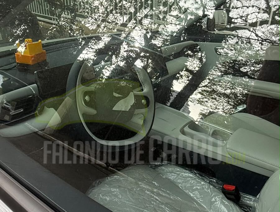 Visão através da janela de um carro novo, com o interior branco e assentos cobertos por plástico. O volante é visível, e a paisagem externa reflete no vidro, mostrando árvores e folhagens. O texto FALANDO DE CARRO está sobreposto na imagem