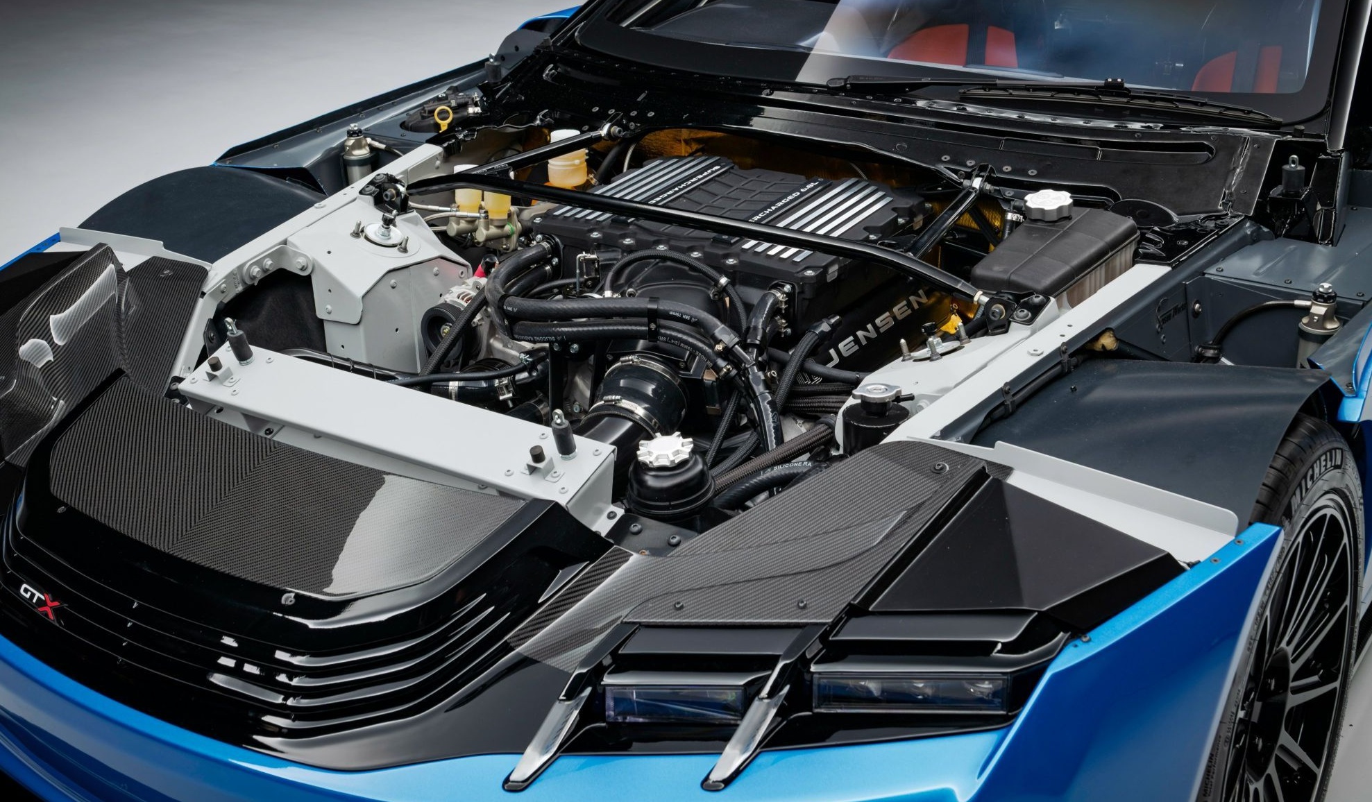 Motor de carro esportivo azul com capô aberto, revelando o motor preto com detalhes prateados e a marca JENSEN, cercado por componentes mecânicos e tubulações. A frente do veículo exibe uma grade preta e elementos de fibra de carbono.
