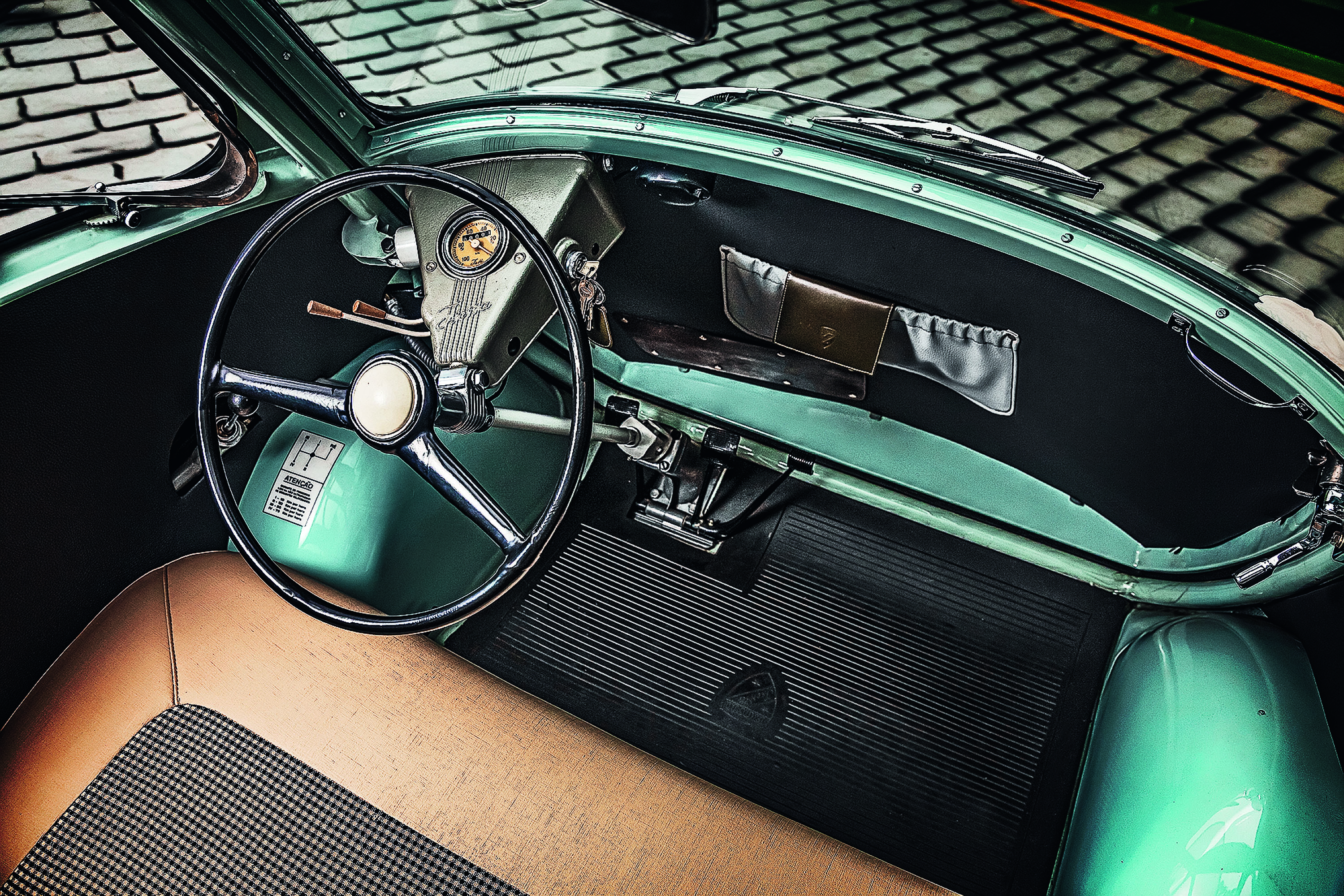 Interior de um carro antigo verde-água, com volante preto e cromado, painel minimalista com velocímetro e porta-luvas embutido. O banco é marrom claro com estampa xadrez, e o assoalho tem tapete preto emborrachado.