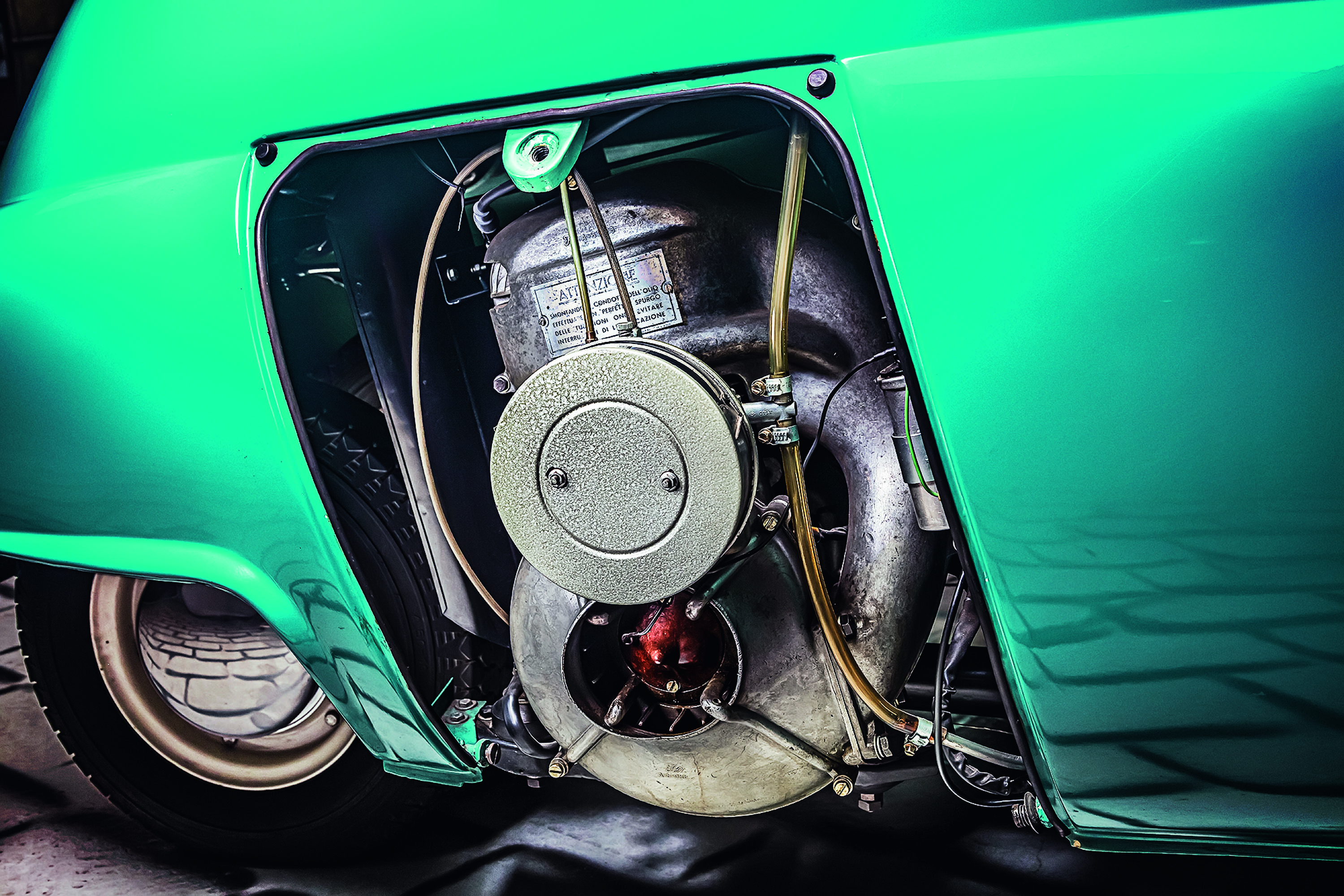 Motor exposto de um carro antigo verde-água, com componentes metálicos prateados e tubulações. A roda traseira é visível na parte inferior esquerda