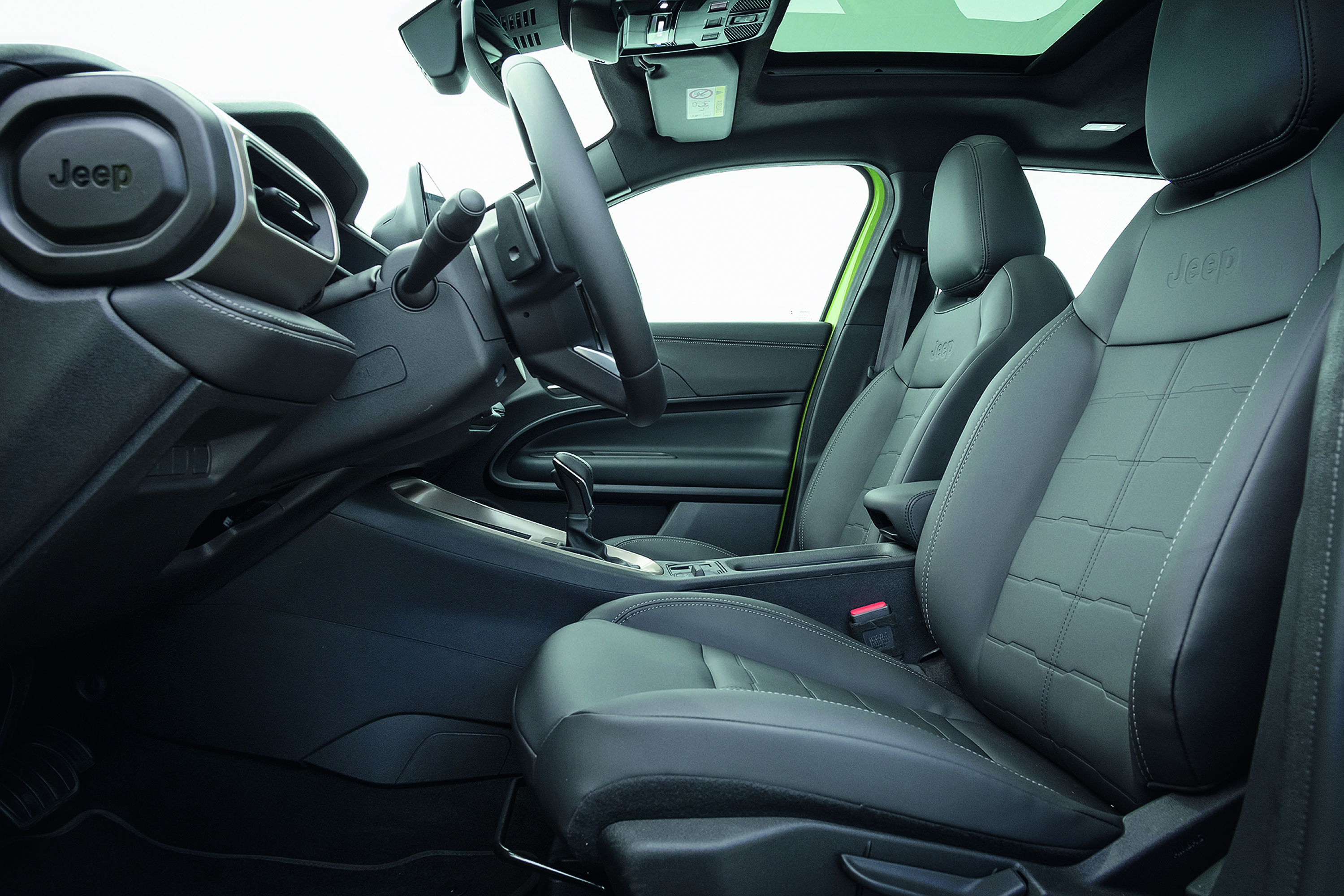 Interior de um carro Jeep, mostrando o banco do motorista e passageiro dianteiro em couro cinza escuro com costuras brancas e detalhes quadriculados. O volante e o painel são pretos, com o logo Jeep no canto superior esquerdo. O teto solar panorâmico e as colunas verdes claras são visíveis, com o câmbio automático no console central