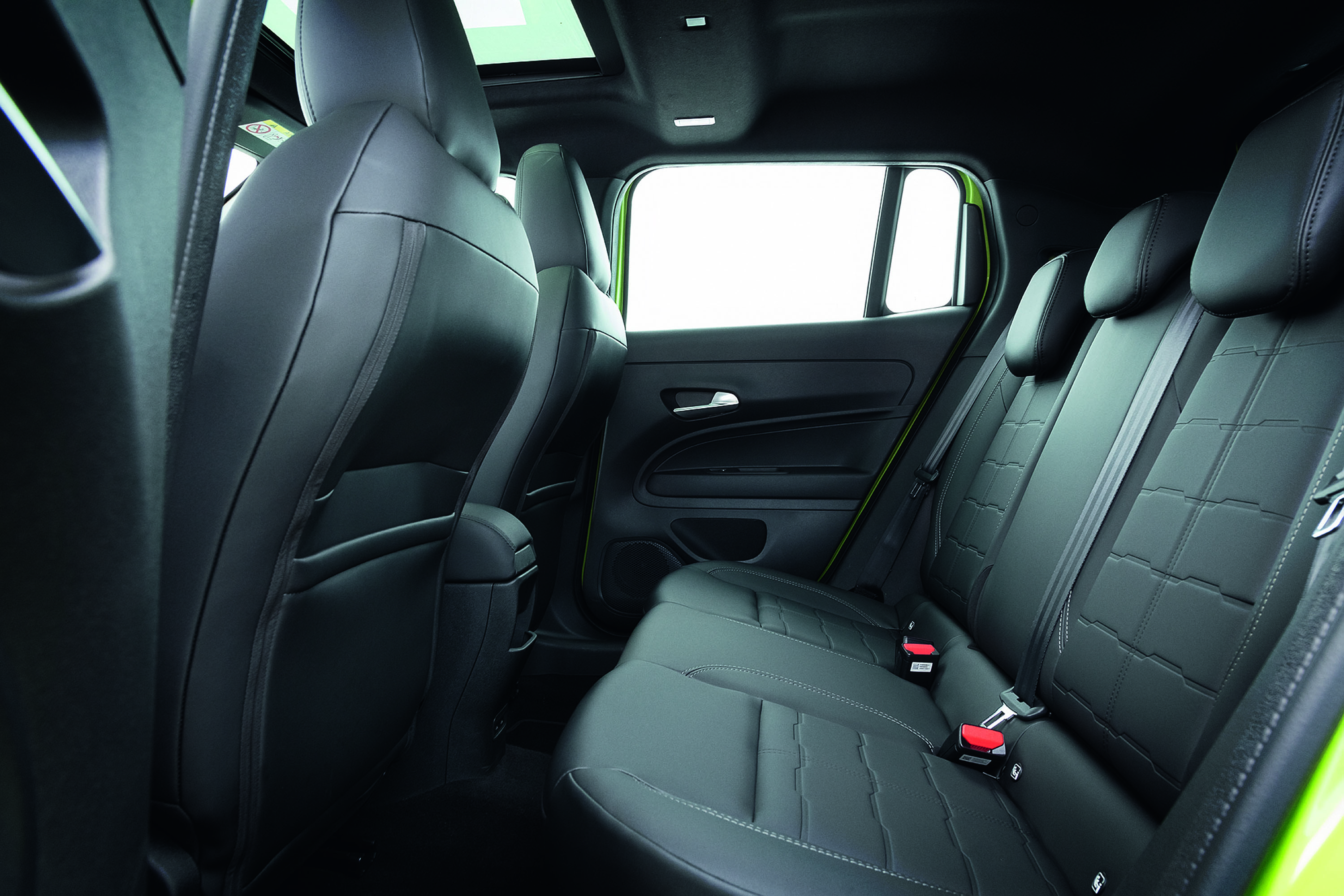 Interior de carro com bancos traseiros de couro preto, com costuras em relevo e cintos de segurança vermelhos. A porta lateral tem acabamento escuro e a janela está aberta para um fundo branco. O teto solar está visível