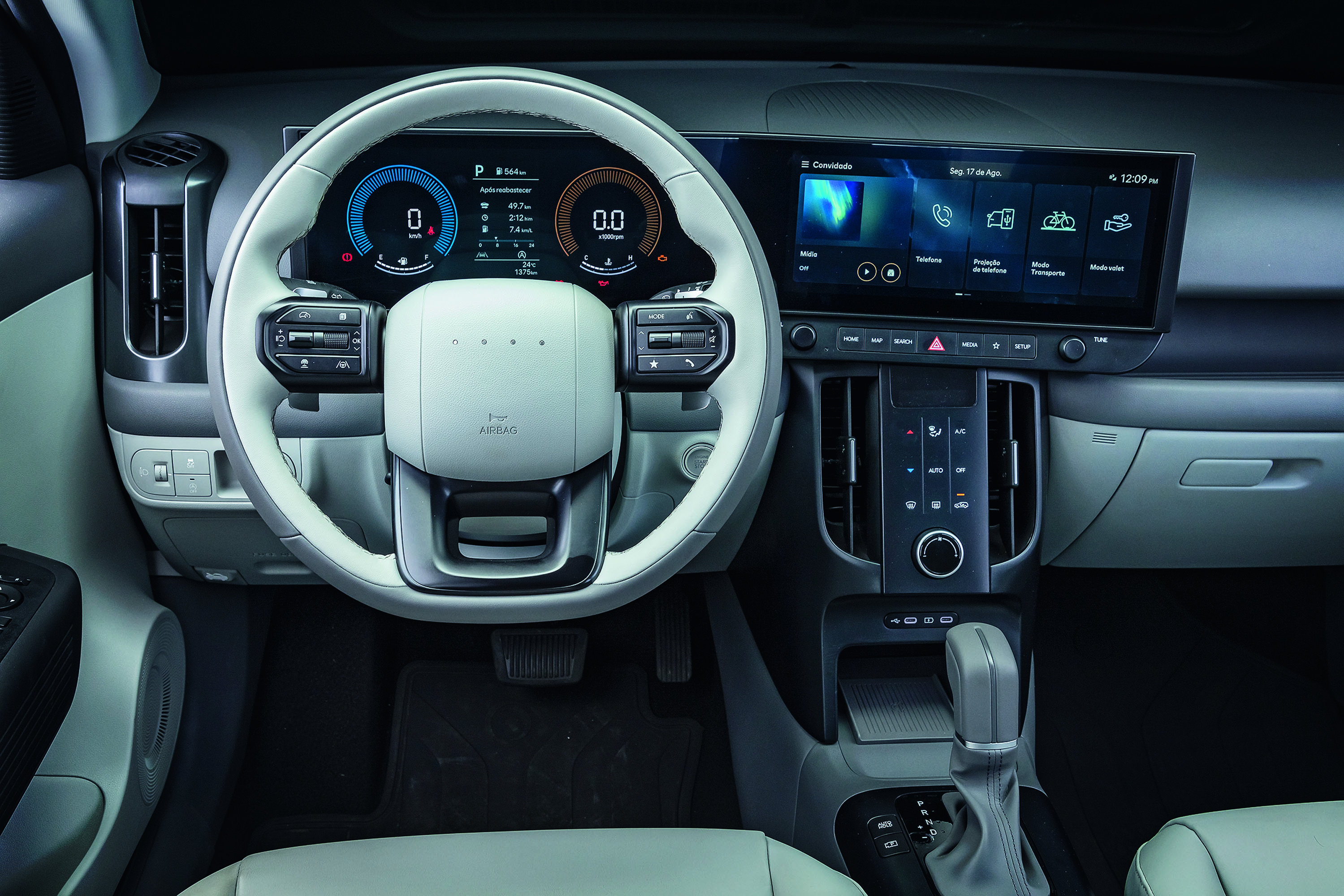 Interior de um carro moderno, com volante branco e cinza, painel digital exibindo velocidade zero e conta-giros. Uma tela multimídia grande mostra ícones de navegação e conectividade. O console central tem saídas de ar verticais e controles de climatização. O câmbio automático está à direita, com detalhes em cinza e preto. O ambiente é clean e tecnológico