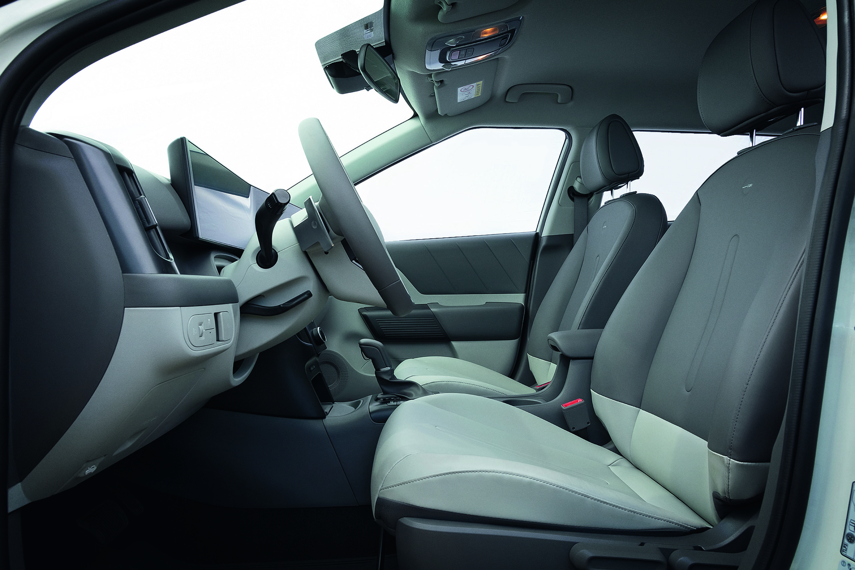 Interior de um carro com bancos dianteiros em dois tons de cinza, claro e escuro. Volante e painel em cinza claro, com tela digital e espelho retrovisor visíveis. O teto é claro e as janelas mostram um fundo branco